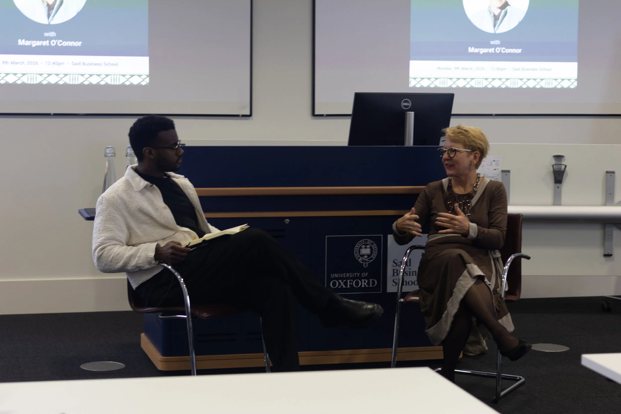 A man and a woman are engaged in a conversation the University of Oxford. The young man is sitting with a notebook, while the woman is speaking and gesturing with her hands. There are two screens behind them displaying a presentation with the name Ma