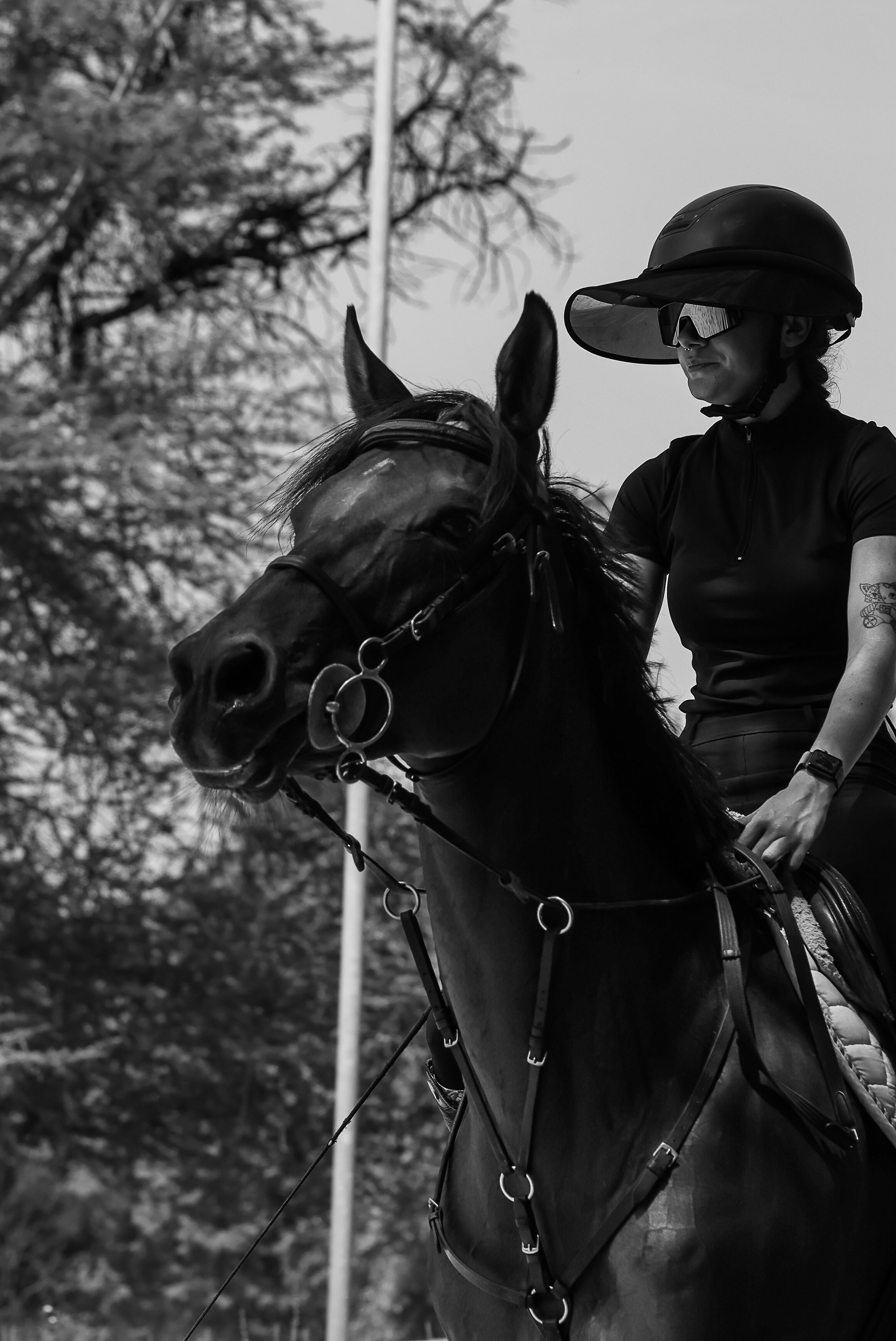 A woman wearing a helmet and sports sunglasses, riding her horse outdoors with trees in the background. Black and White.