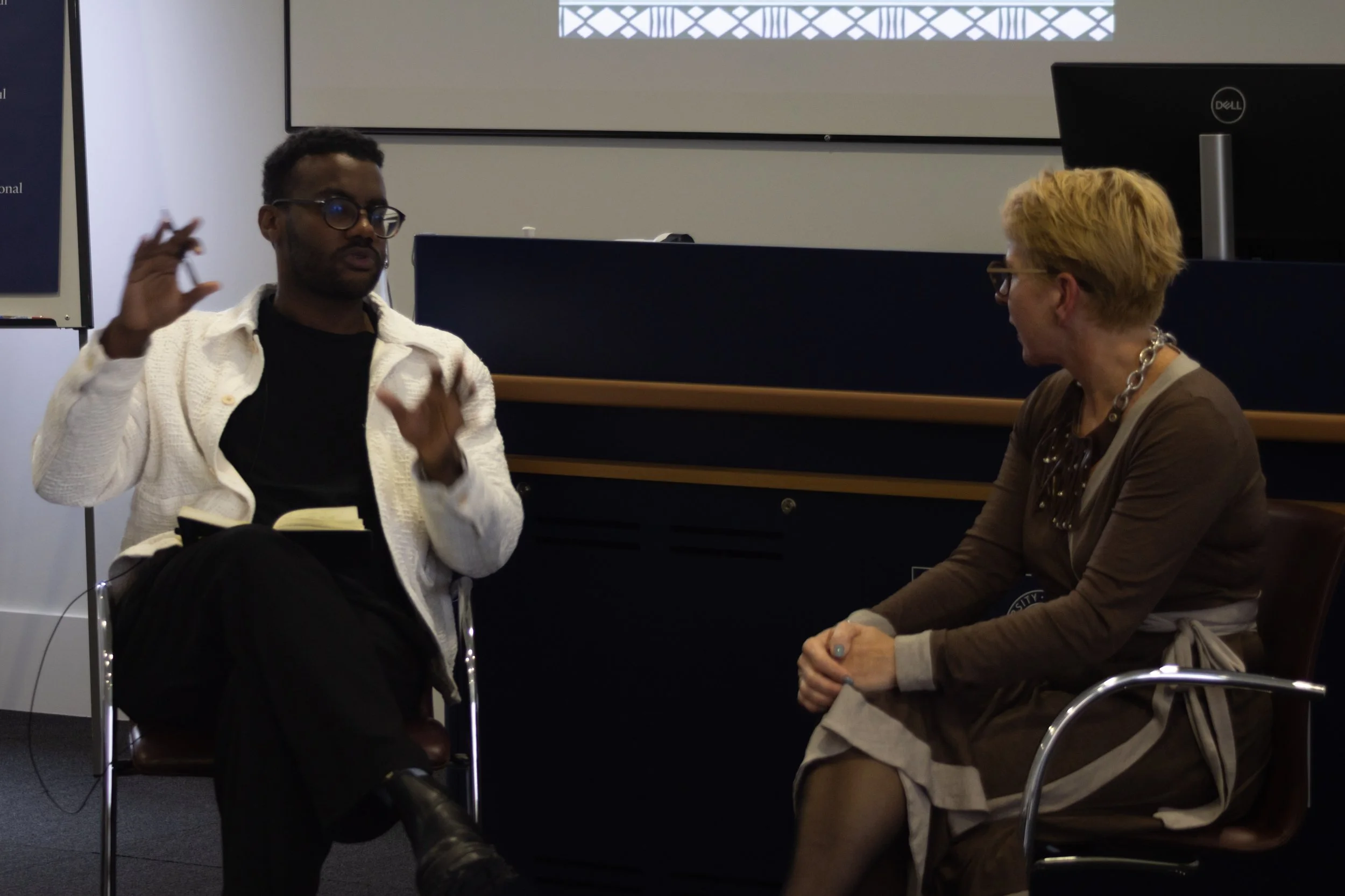 Two women sit facing each other in discussion, one with short blond hair, glasses, and a brown cardigan, and the other with short dark hair, glasses, and a white jacket, inside a room with a projector screen in the background.