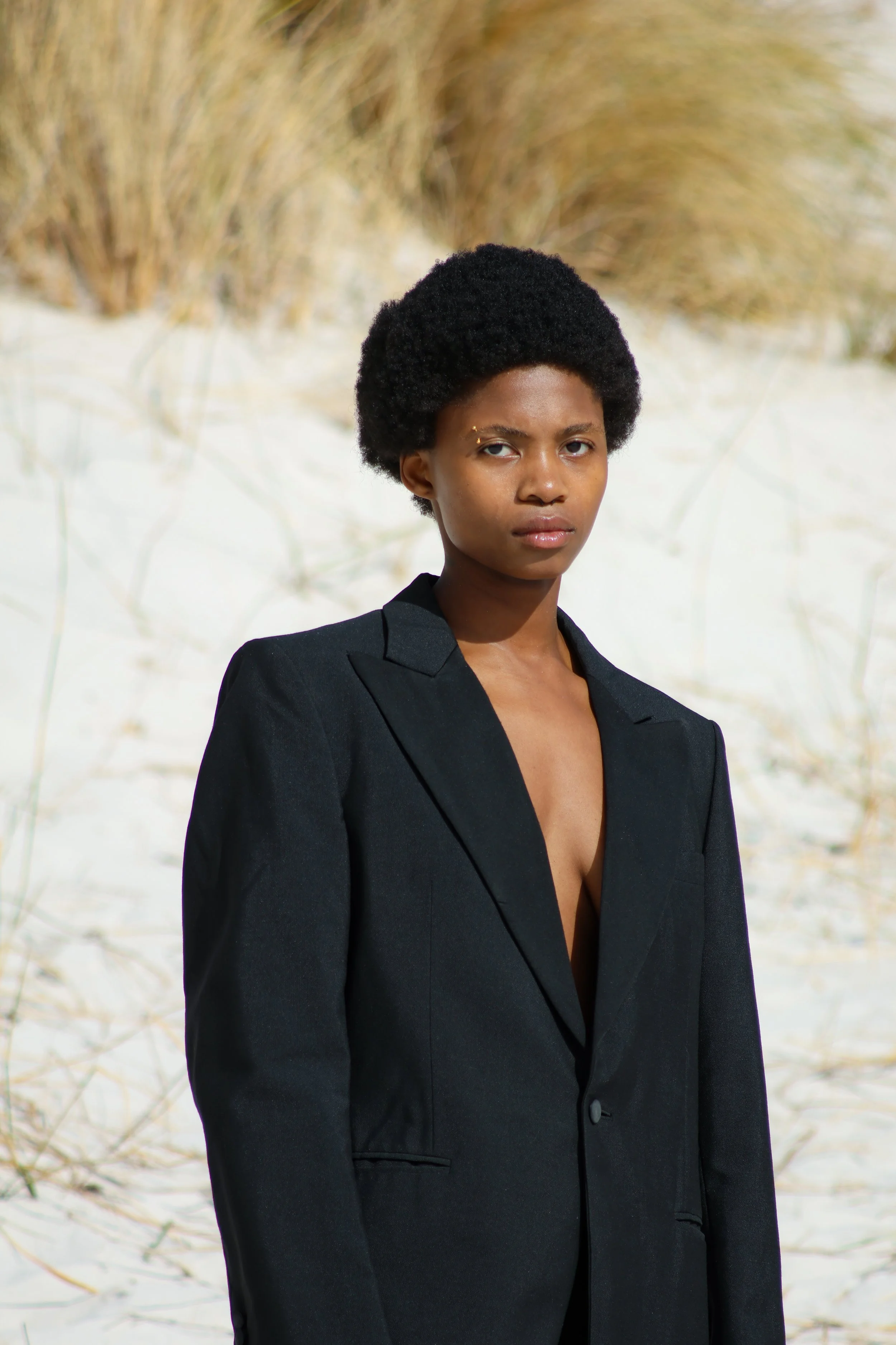 A woman standing on a sandy beach with beach grass in the background, wearing a black blazer with no shirt underneath, and looking directly at the camera with a serious expression. Noordhoek beach, Cape Town.