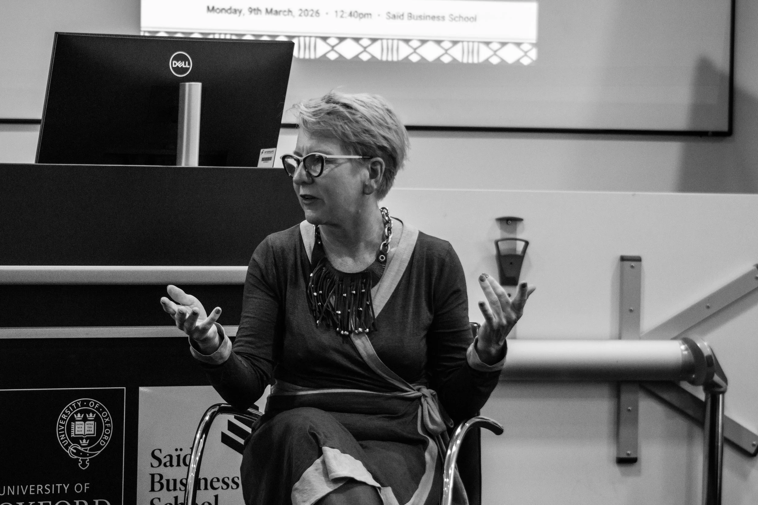 A woman with short hair, glasses, and a patterned dress, speaking or gesturing while sitting in a chair in front of a computer monitor at a university event.