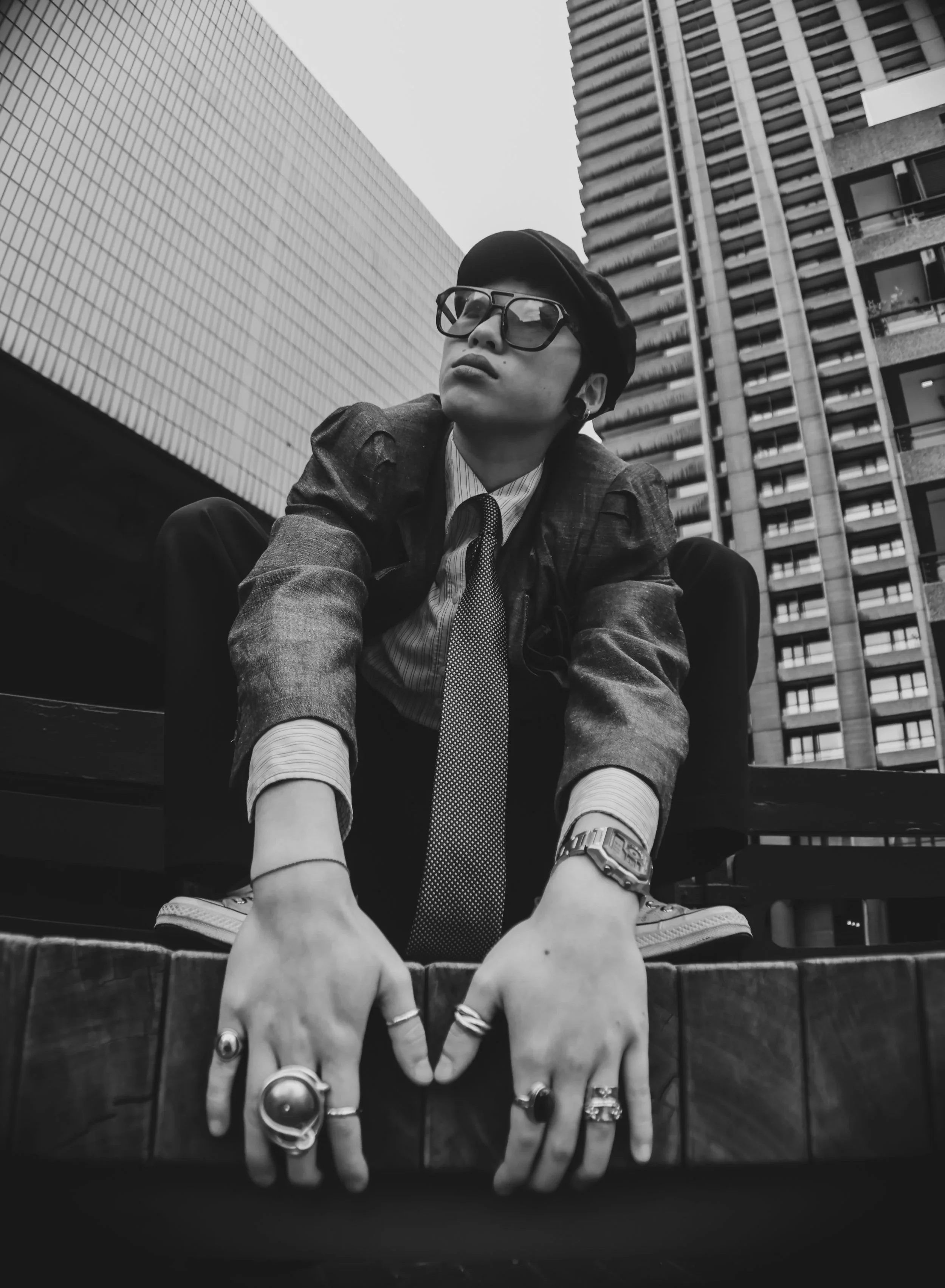 A person wearing glasses, rings, a watch, a tie, and a jacket, crouching with hands on a wooden surface in an urban area with tall buildings in the background, in black and white.