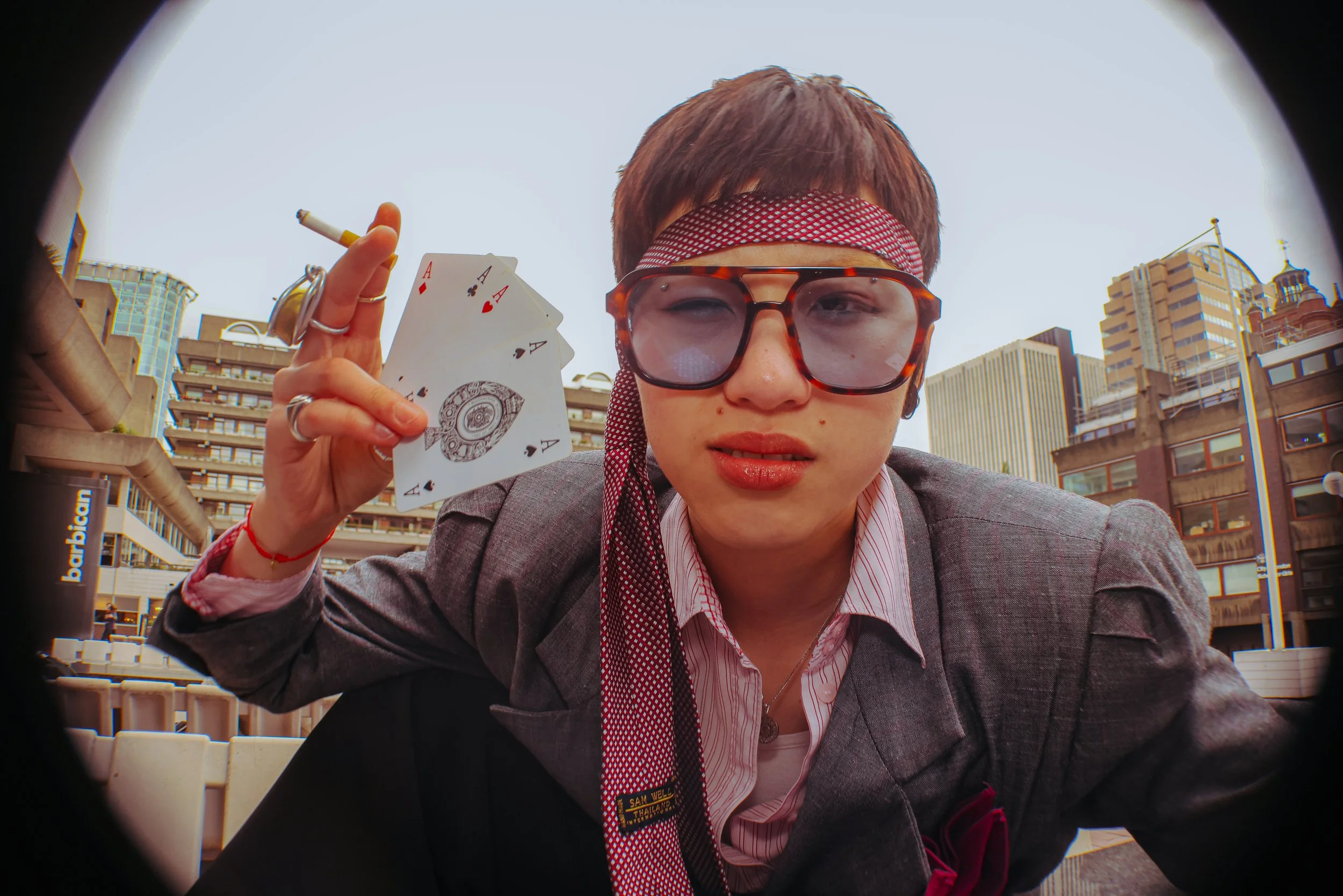 Close-up of a person dressed in a costume with glasses, holding four aces in one hand, with a cityscape in the background.