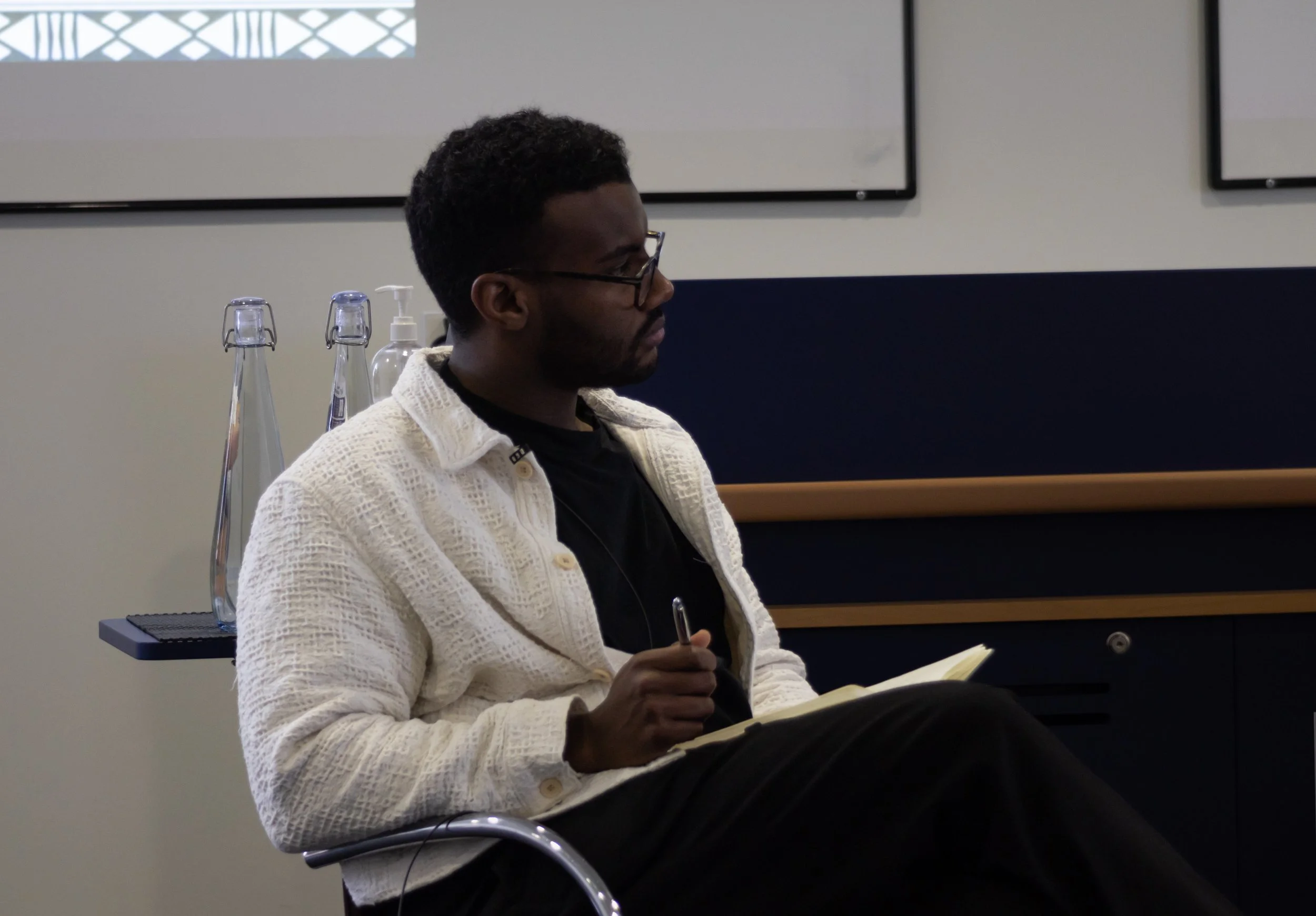 A man sitting in a chair, writing in a notebook, with glasses and a white textured cardigan, in a room with bottles and a cart in the background.