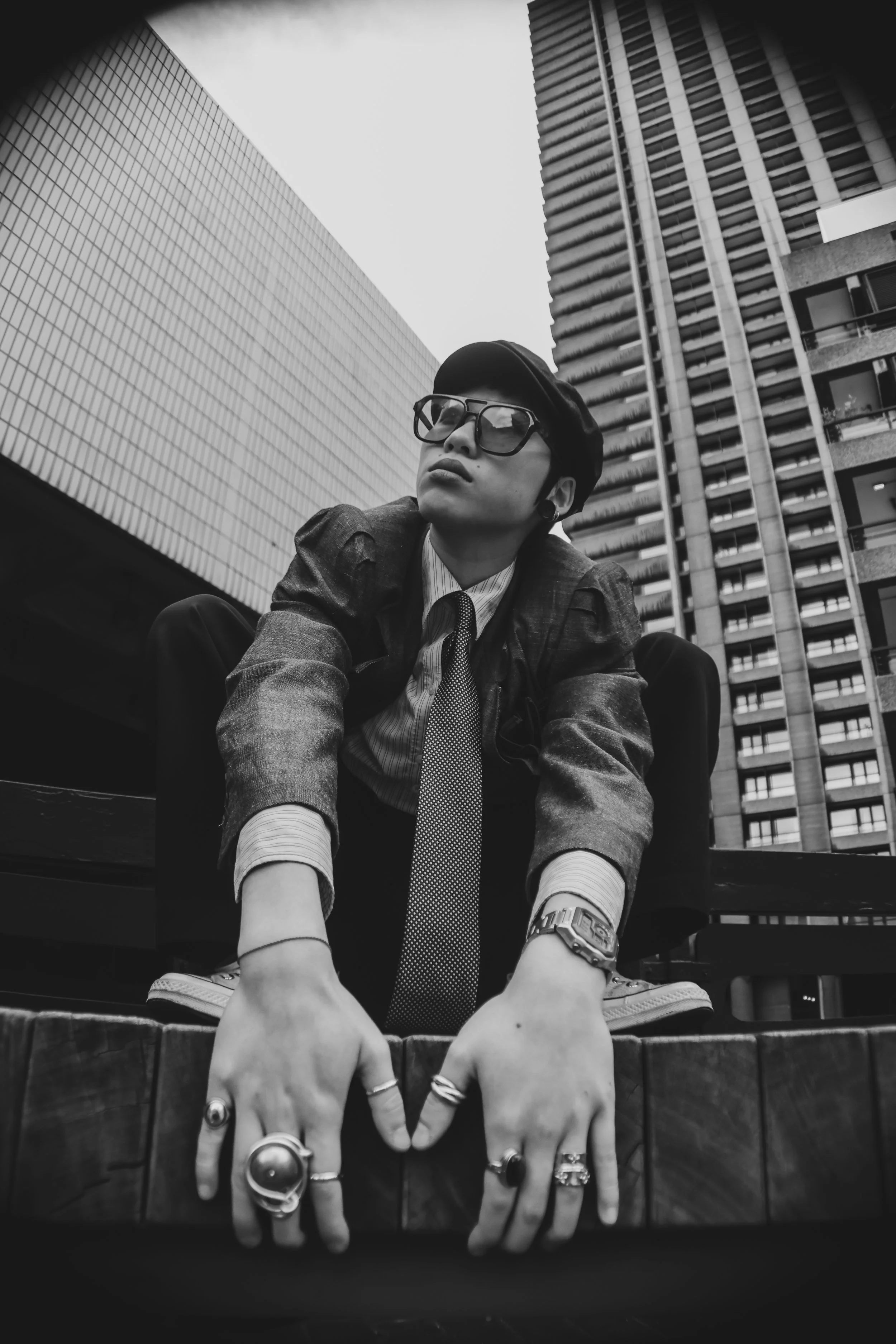 A black and white photo of a person wearing glasses, a hat, a patterned tie, and a watch, crouching on a ledge in an urban setting with modern skyscrapers in the background.