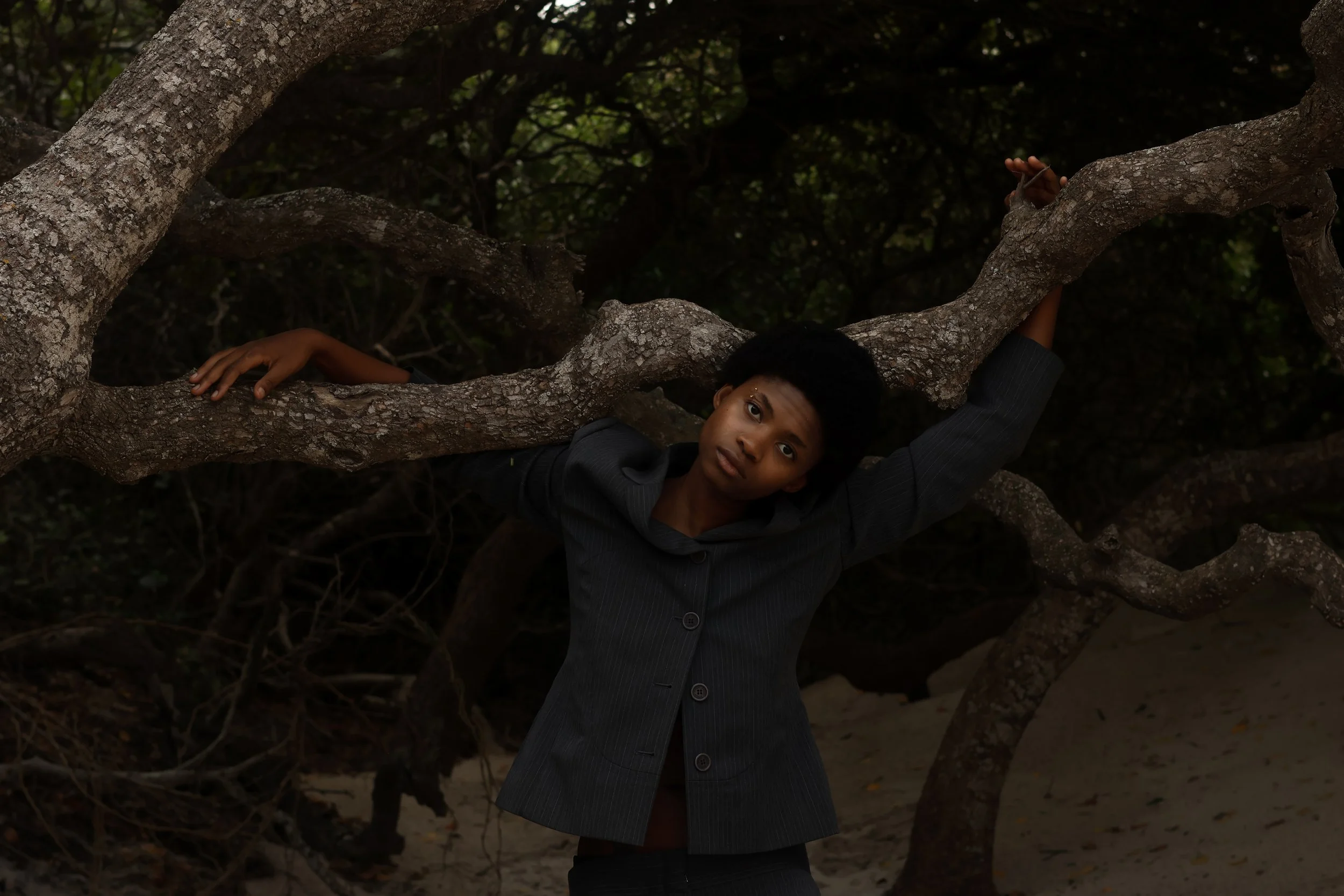 An editorial portrait of a young person with short hair wearing a dark blazer, leaning on a large tree branch in a forested area with sand ground.  Noordhoek beach, Cape Town.