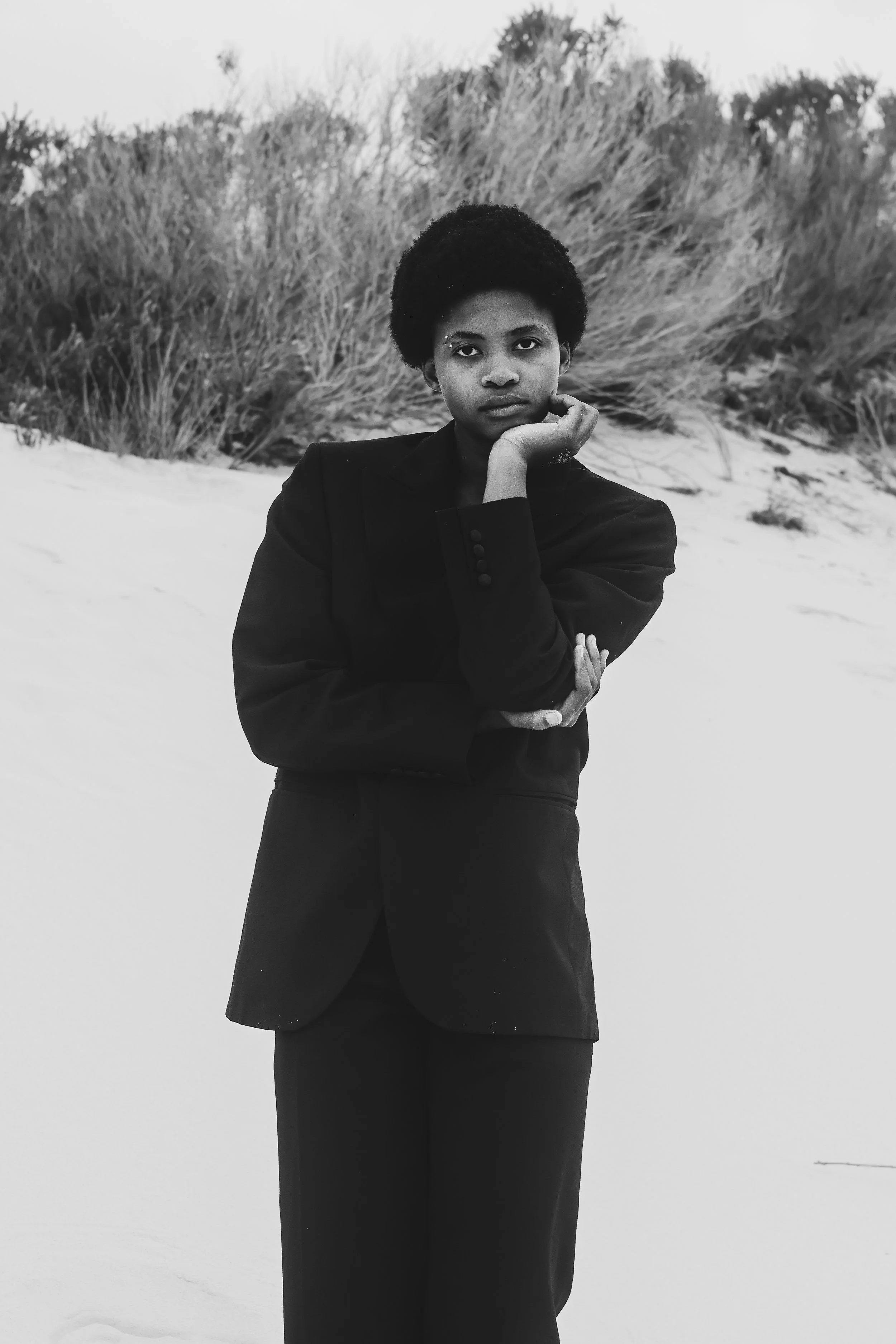 A woman with short curly hair wearing a black blazer and pants standing on sand dunes with tall grass in the background, posing with her hand resting on her face in a black and white photograph. Noordhoek beach, Cape Town.