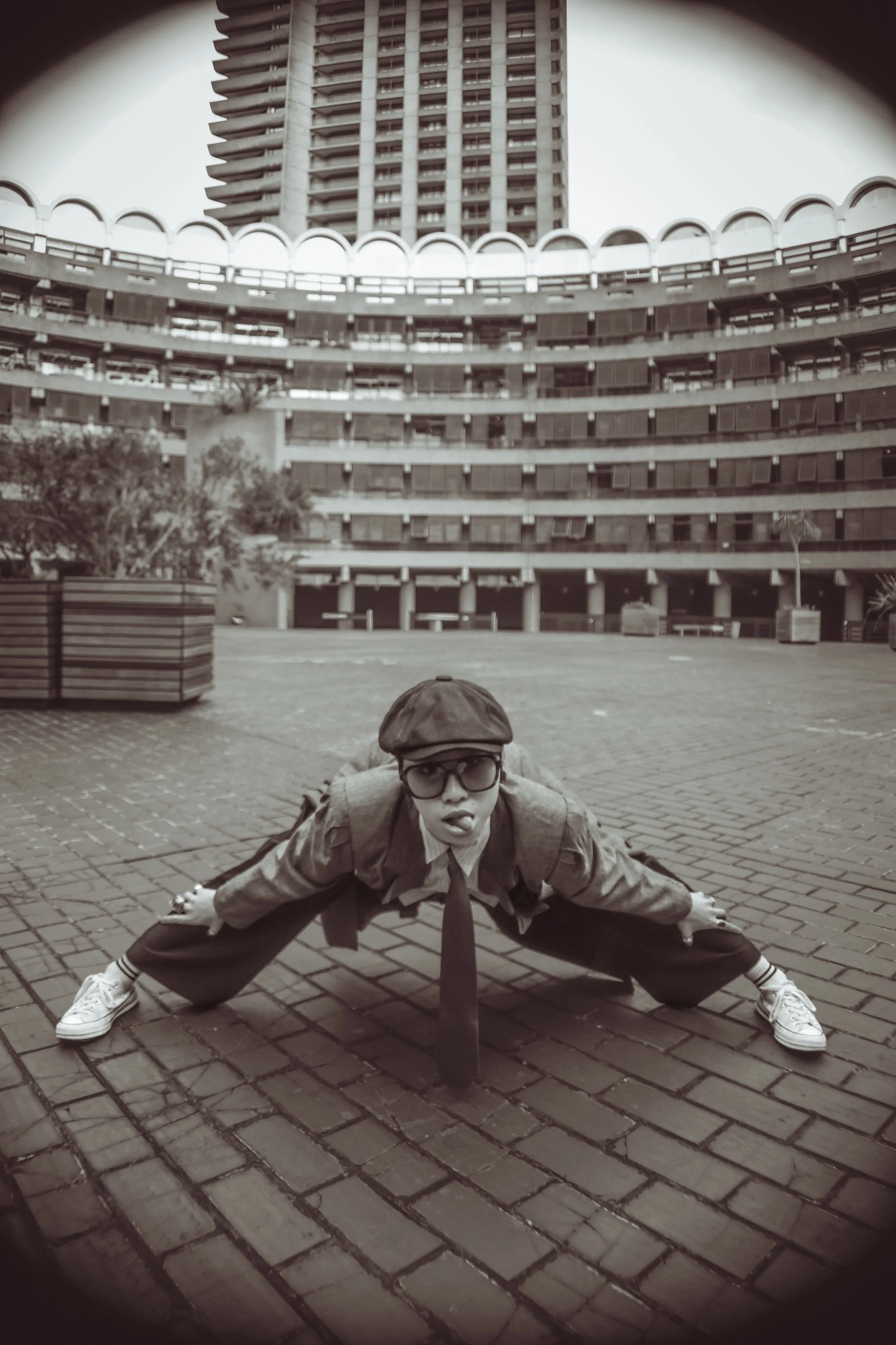 Person in retro style clothing, including cap, glasses, tie, and sneakers, crouching with legs wide apart and hands on the ground in an urban courtyard, with large building in the background.