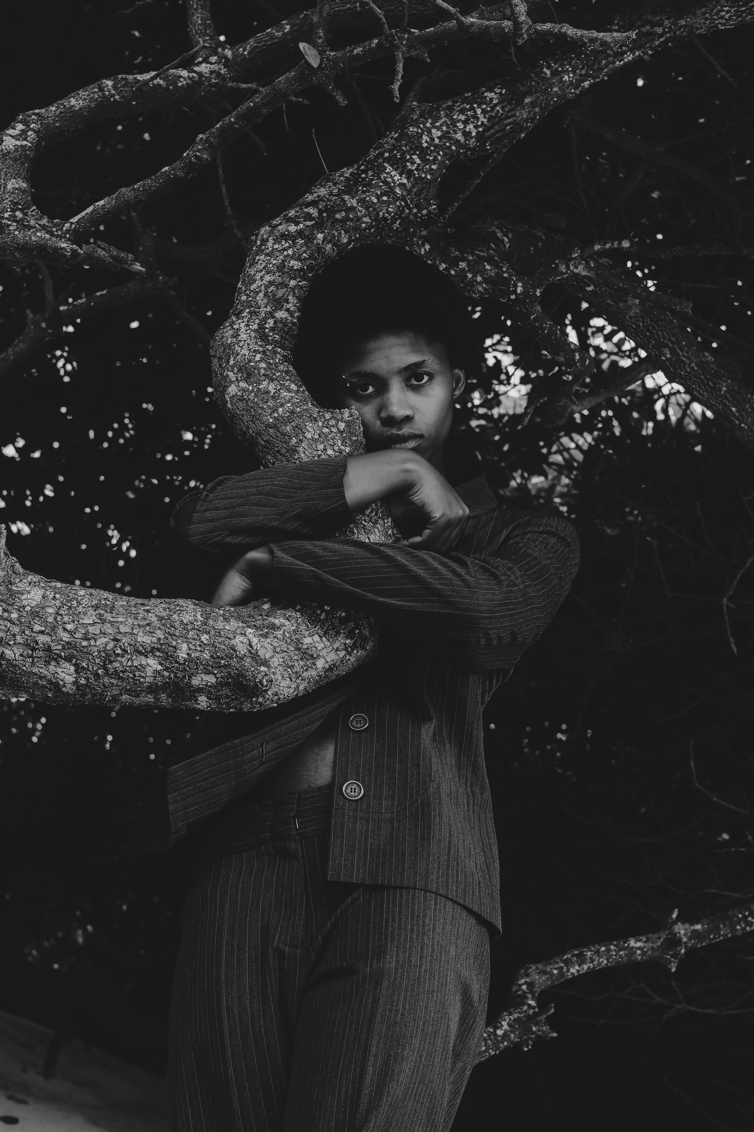 A person leaning against a tree branch, looking directly at the camera, wearing a dark striped suit in a black and white photo. Noordhoek beach, Cape Town.