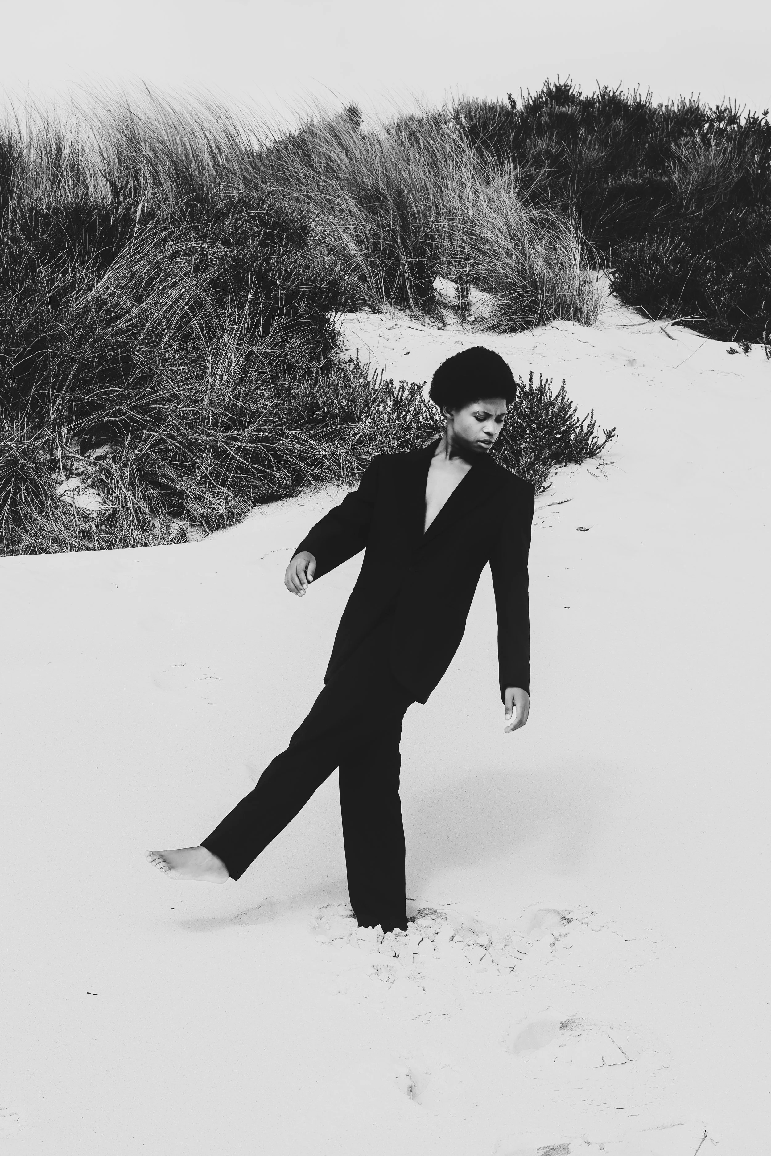 Black and white photo of a woman in a black suit with short curly hair walking barefoot on sandy dunes. Noordhoek beach, Cape Town.
