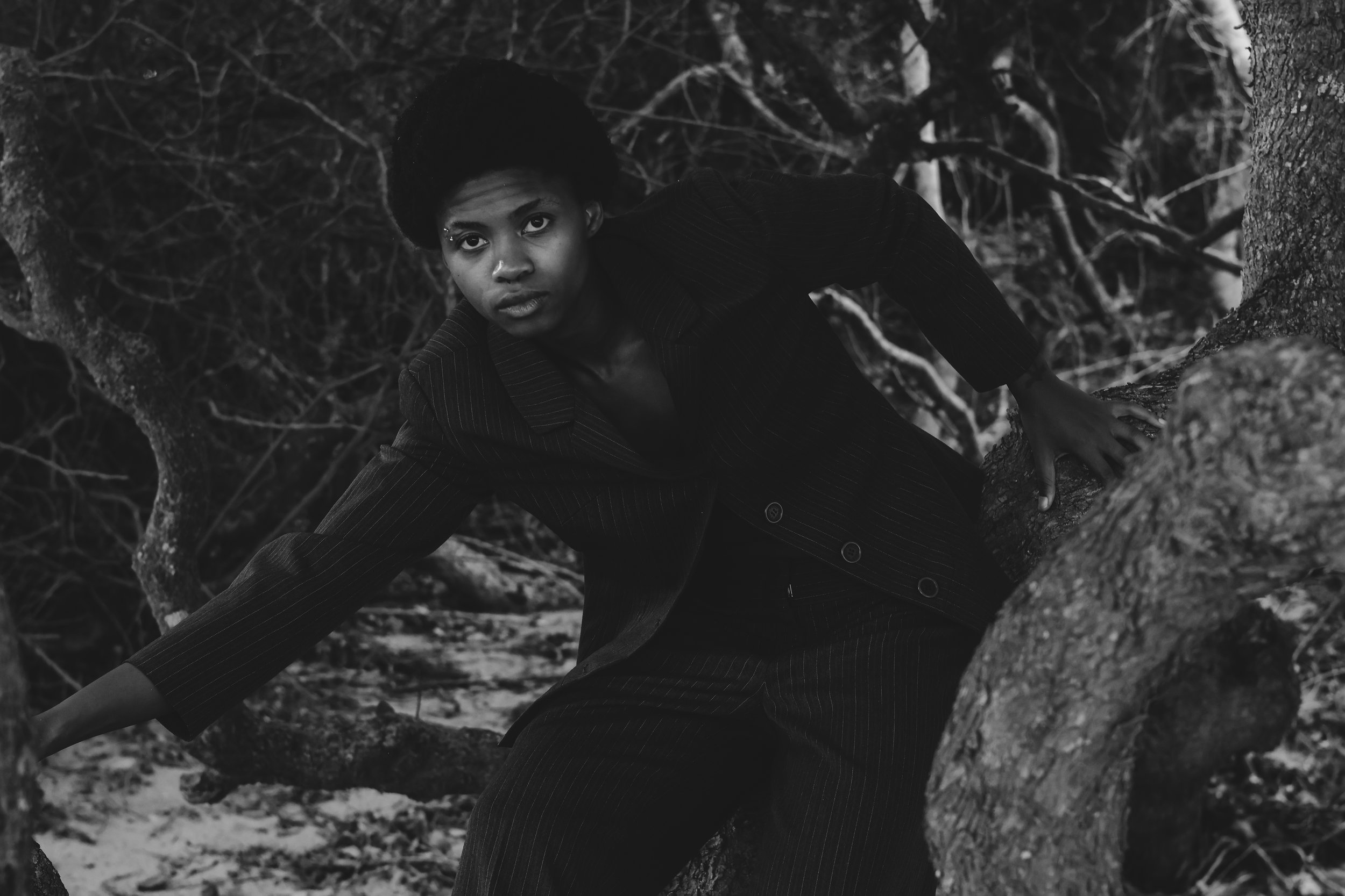 A person with natural hair in a black pinstripe suit crouches on a tree branch among dense tree branches and foliage in a black and white photo. Noordhoek beach, Cape Town.