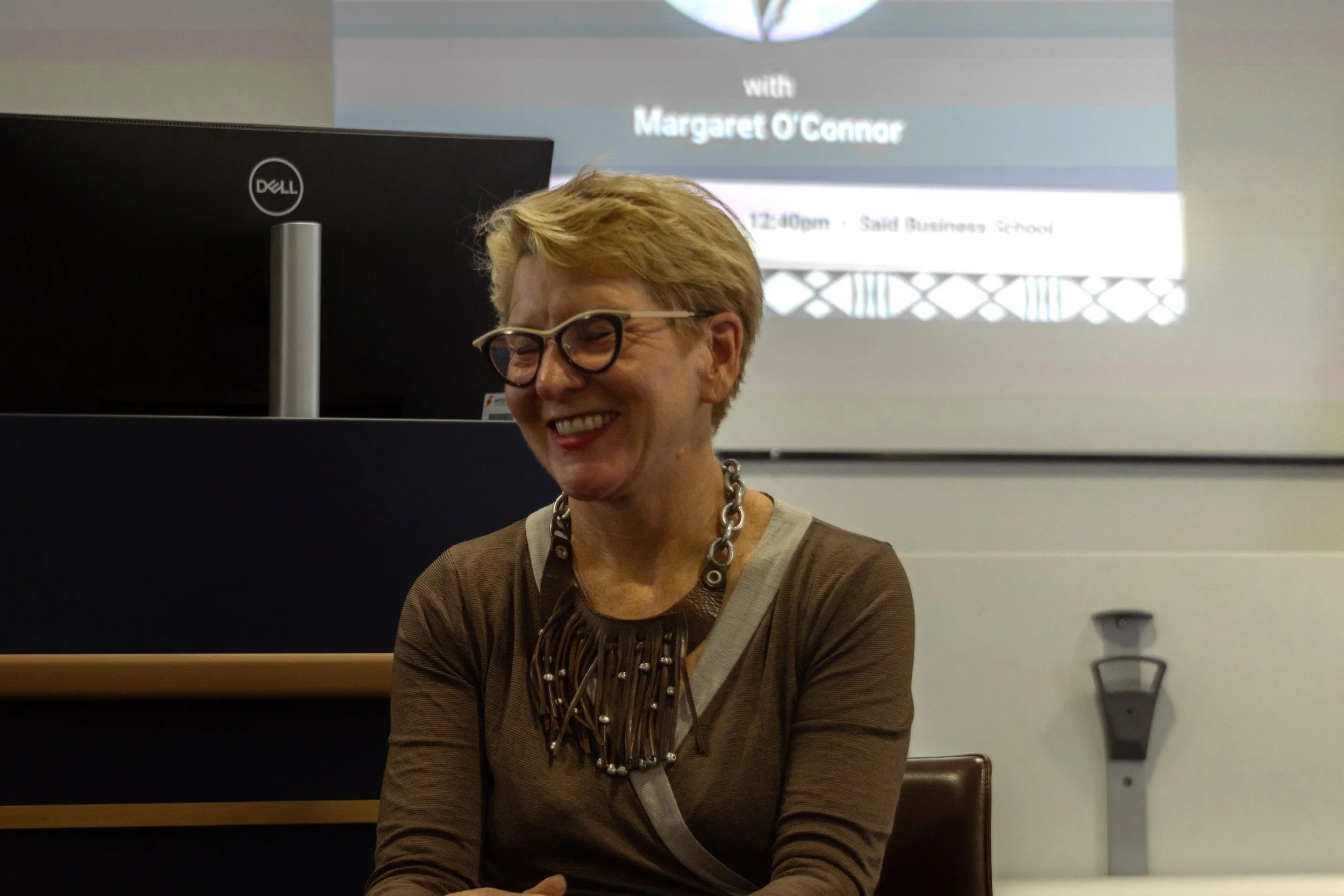 A woman with short blond hair and glasses smiling, sitting in front of a computer monitor with a presentation screen in the background.