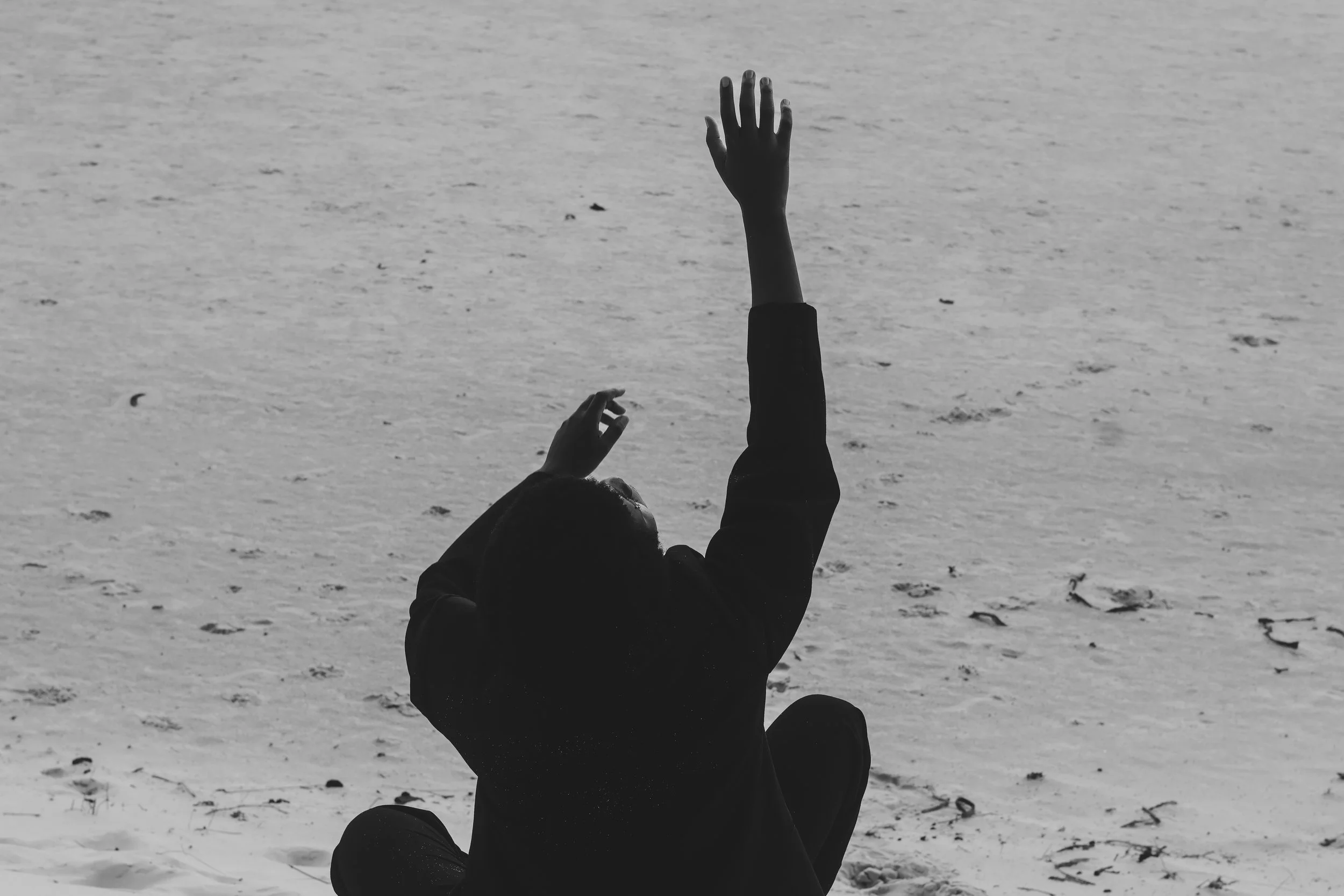 Person sitting on the beach with one arm raised and the other hand near their head, facing away from the camera.