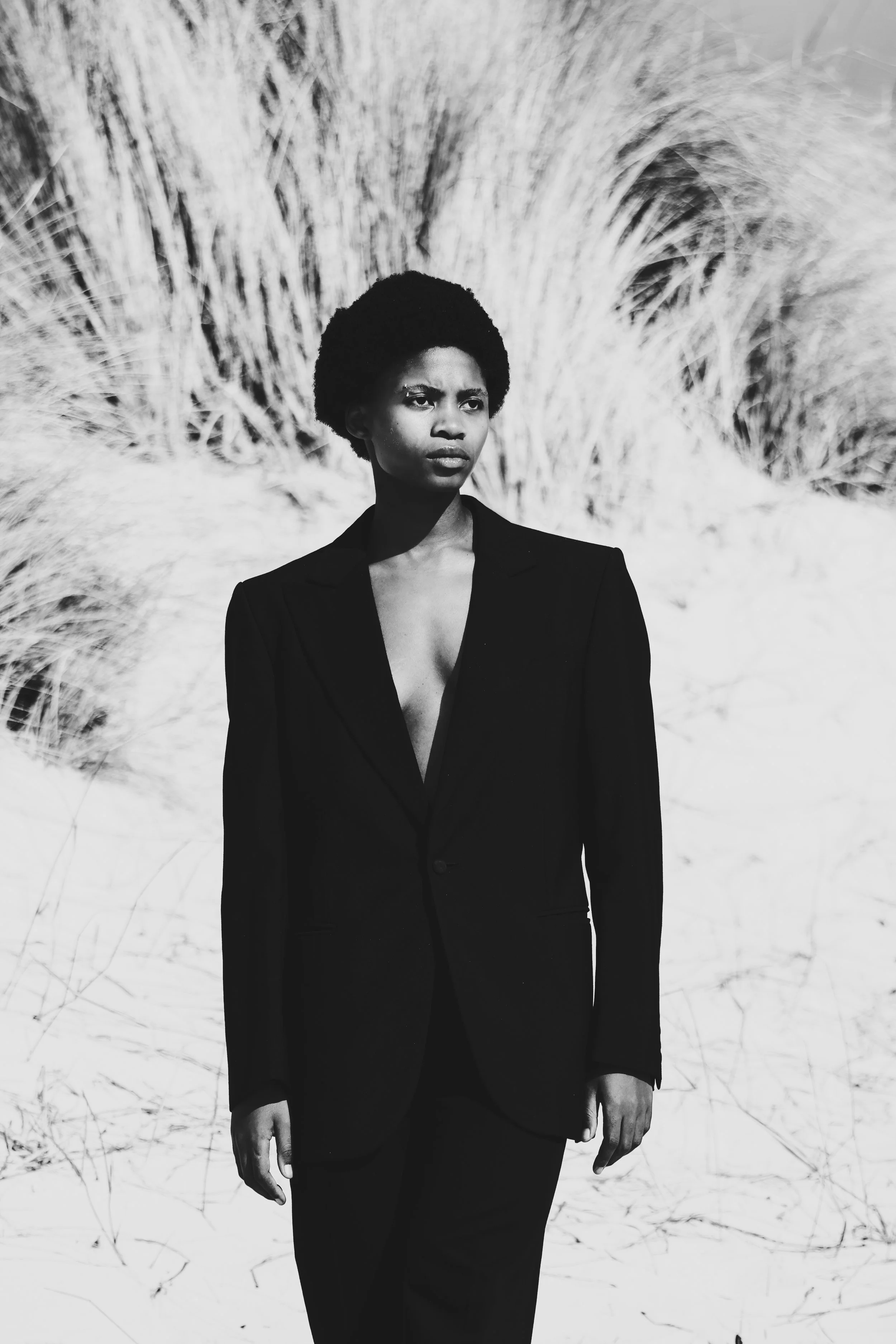 A woman in a black suit standing in a sandy outdoor area with tall grass in the background. Noordhoek beach, Cape Town.