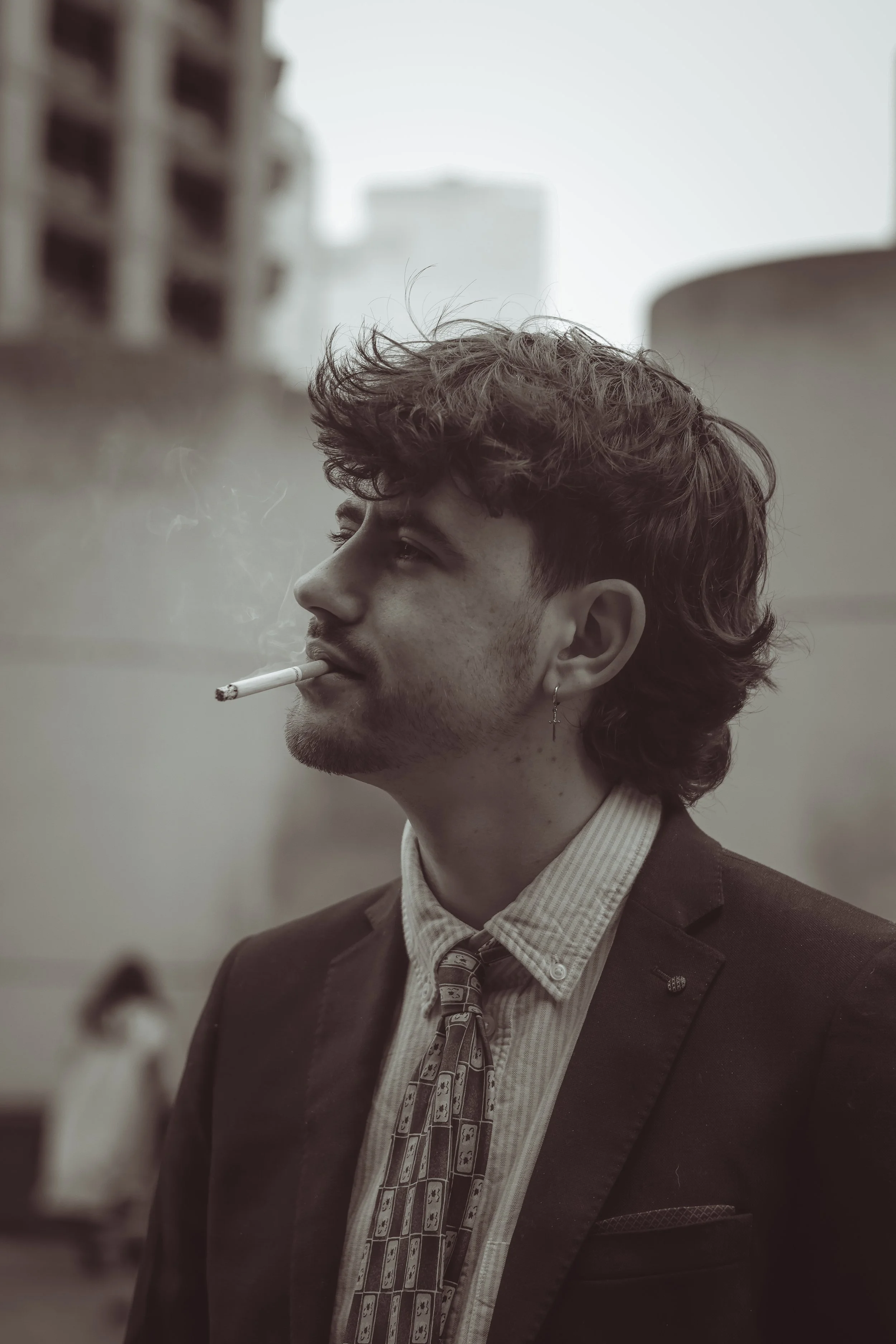 A young man with curly hair and earrings, dressed in a suit and tie, is smoking a cigarette in an urban outdoor setting.