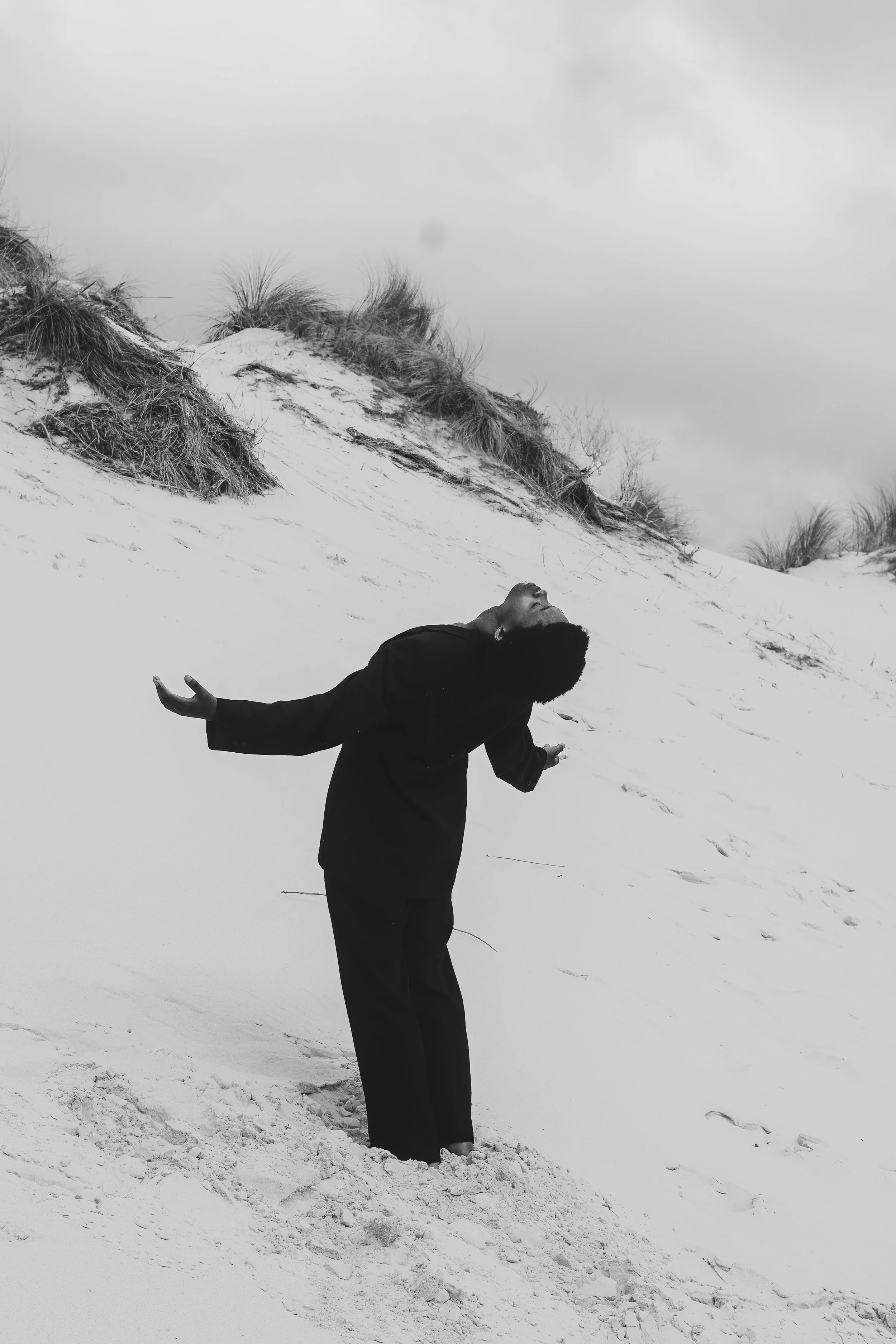 A woman standing on a sandy beach with dunes and grass, looking upward with her arms outstretched and head tilted back. Noordhoek beach, Cape Town.