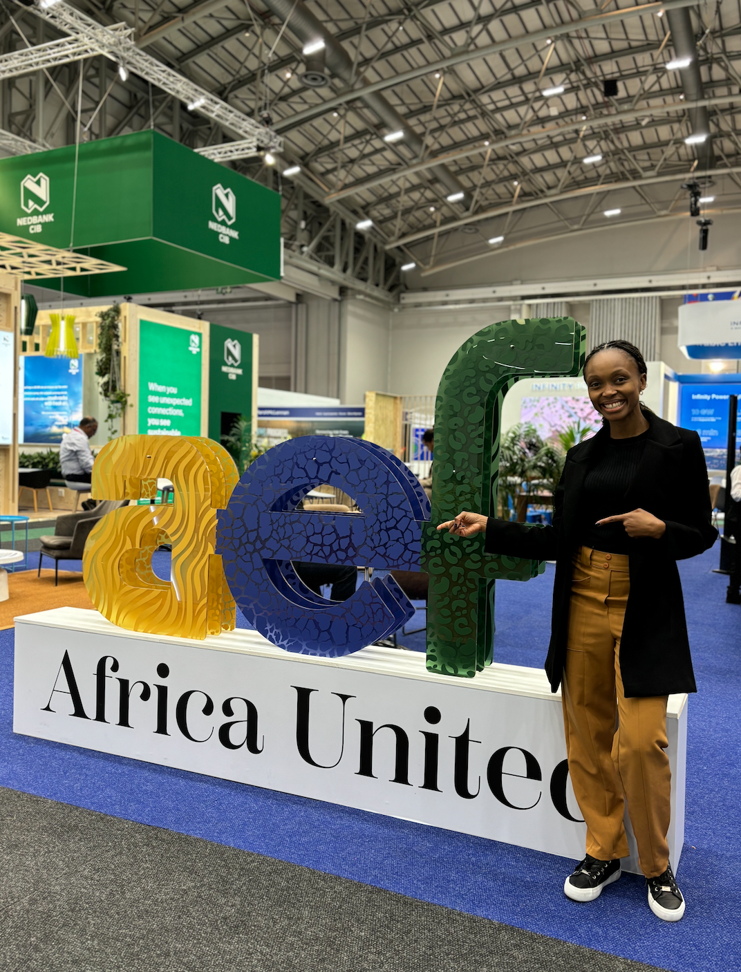 A woman standing next to a large sign that says "Africa United" at an indoor conference. The woman is smiling and pointing towards the sign that says 'AEF'.