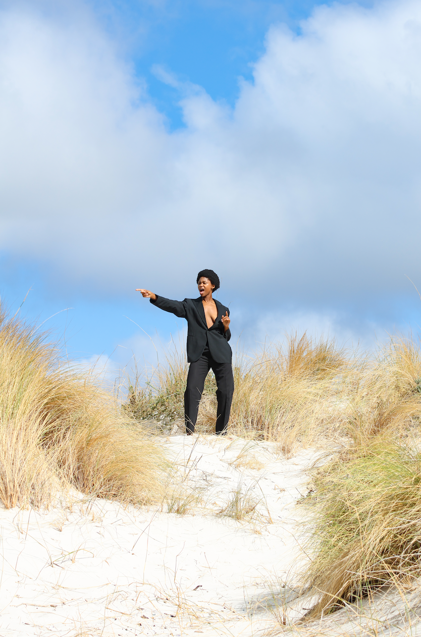 A woman in a black suit standing on a sandy dune with tall grass, pointing and speaking with a cloudy sky in the background. Noordhoek beach, Cape Town.