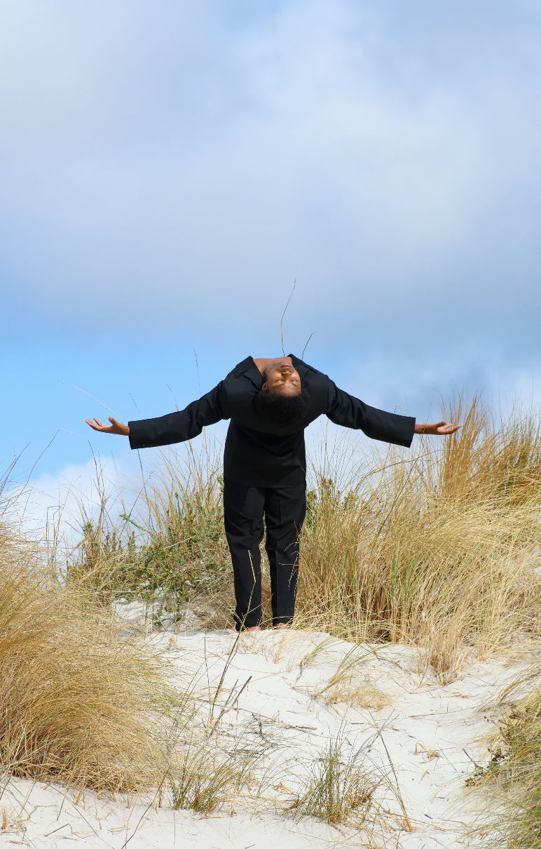 A person in a black suit with arms stretched out, bent forward at the waist, standing on sandy dunes with tall grass, against a partly cloudy sky. Noordhoek beach, Cape Town.