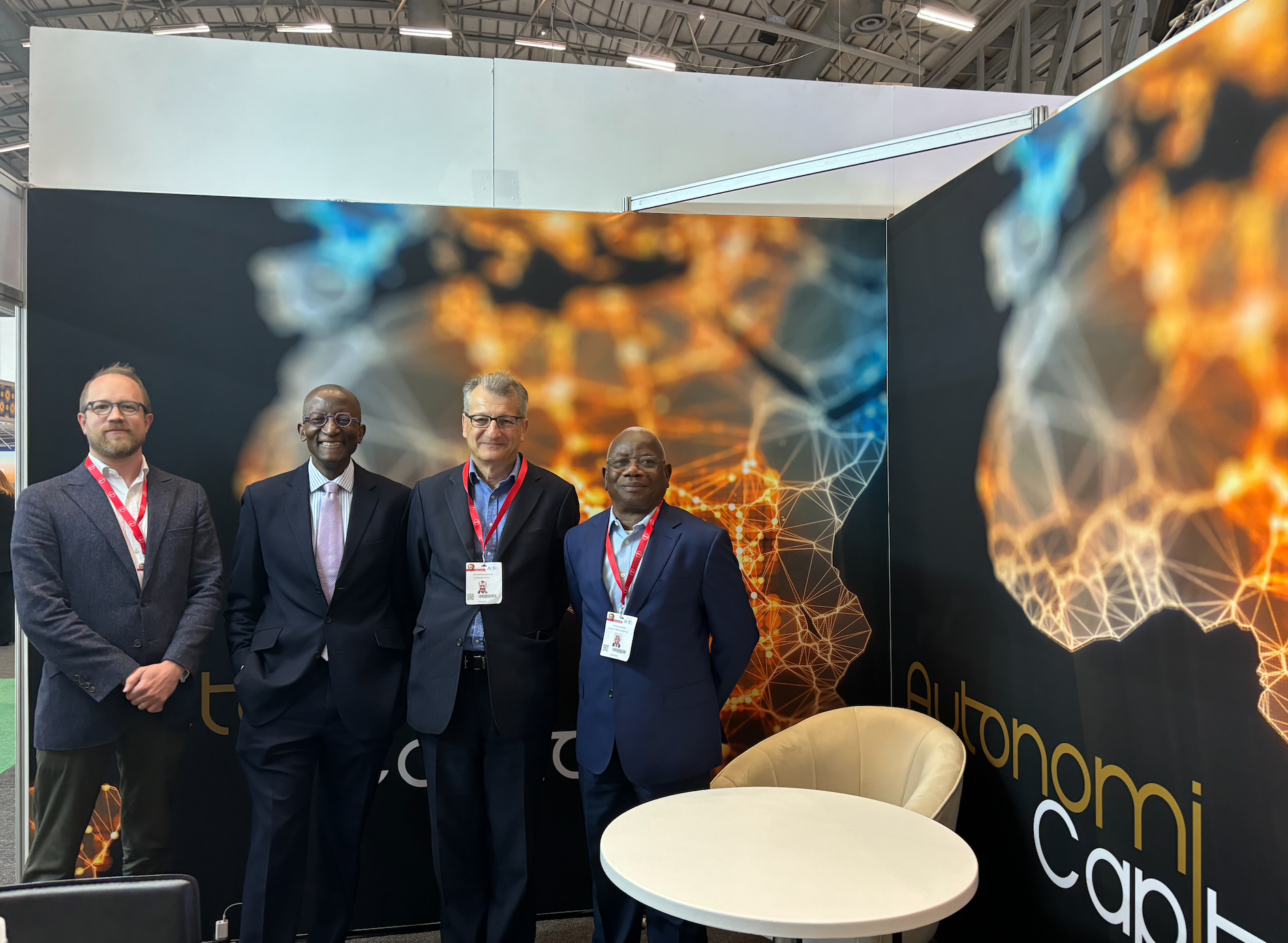 Four men in business attire standing together at a conference booth with a digital-themed backdrop showing the map of the African Continent, with vivid interconnected orange and blue lines and nodes.