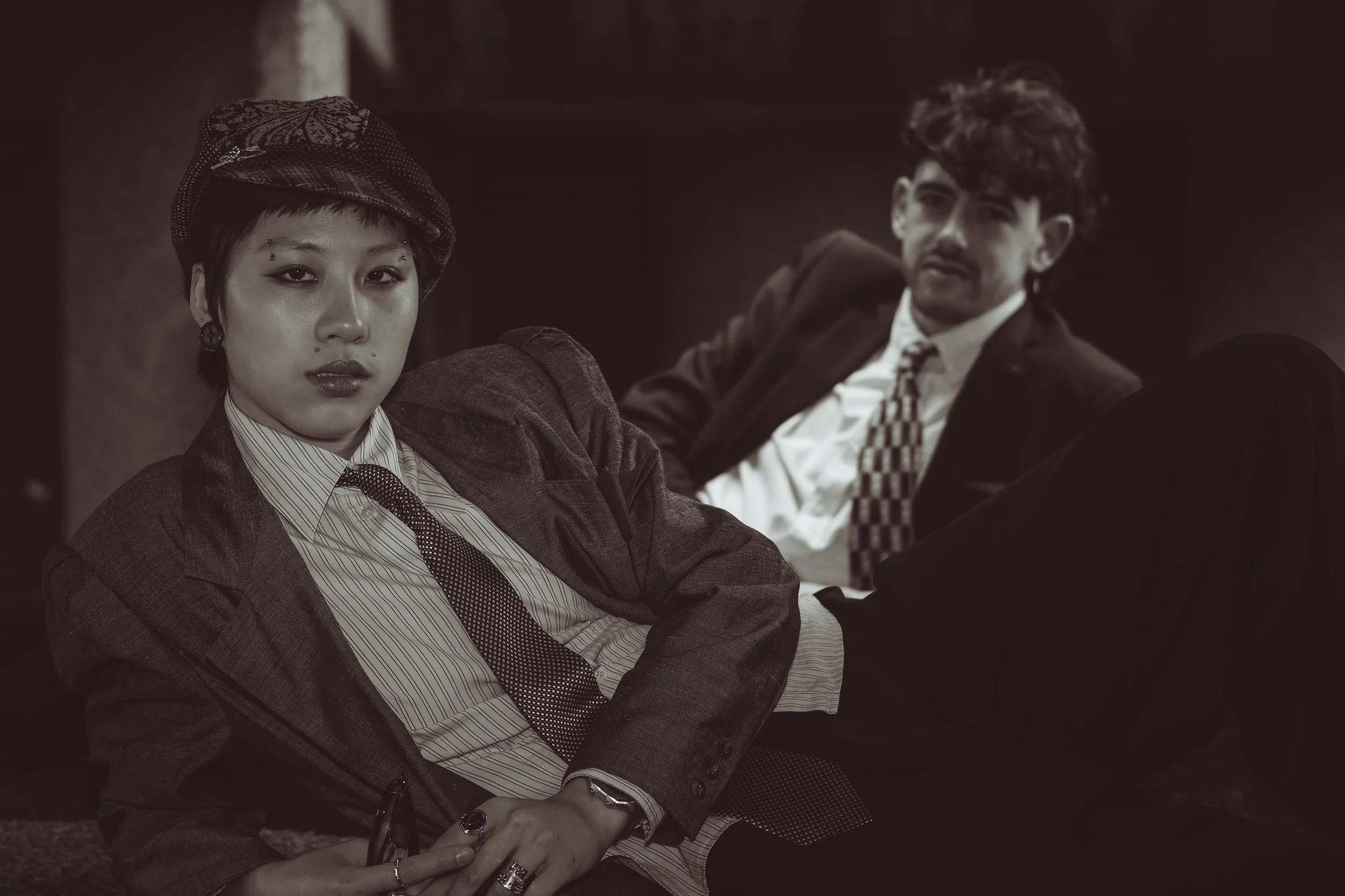 A black-and-white photo of a woman lying on the floor with her head propped up, wearing a plaid blazer, a striped shirt, a tie, and a beret, with earrings and facial piercings. A man in a suit with a patterned tie is sitting behind her, slightly blurred, with an intense expression.