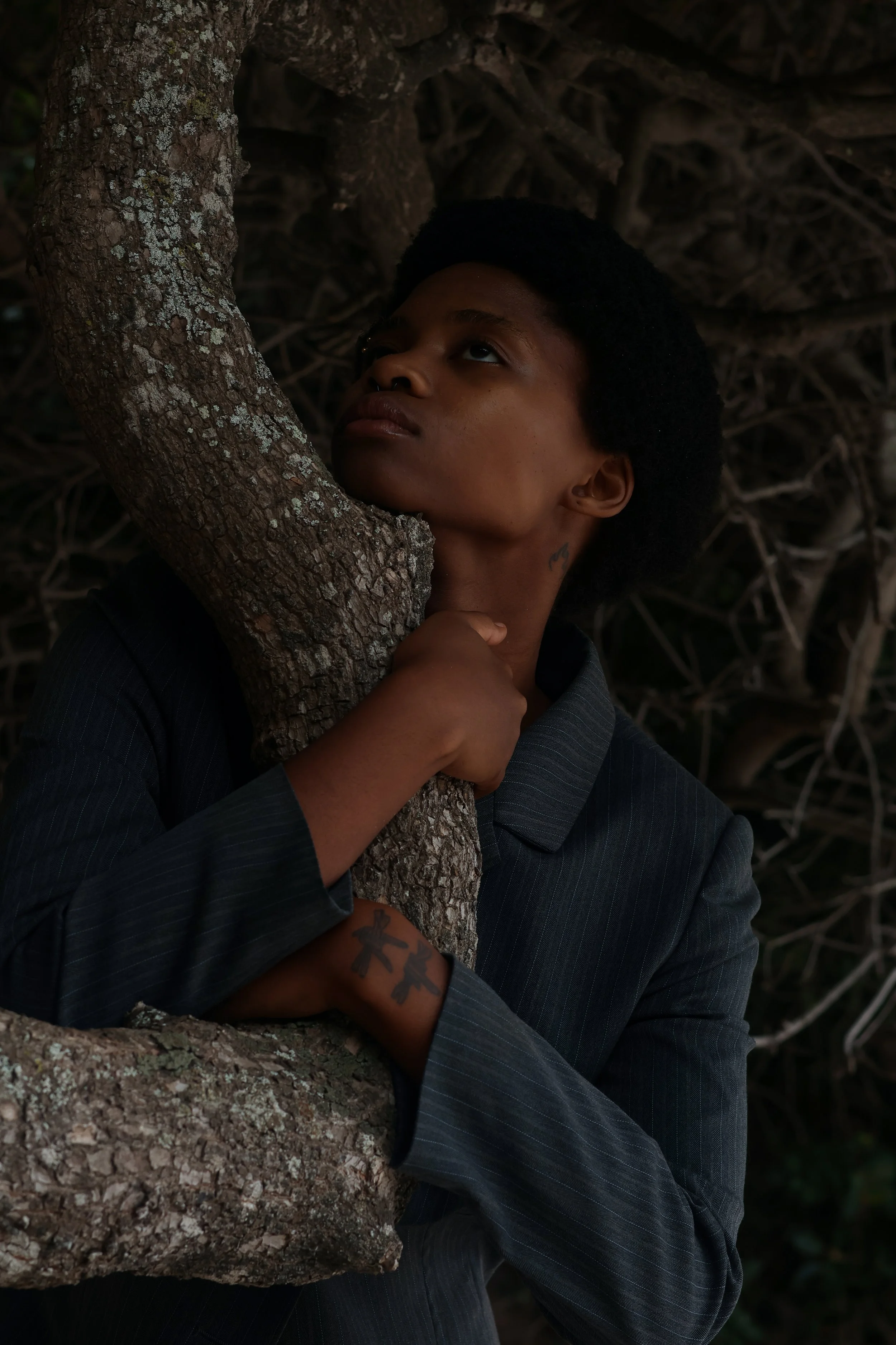 A portrait of a young person holding a tree trunk, with an intense, contemplative expression, wearing a dark pinstripe jacket, with tattoos on their hand and neck, dark background of intertwined branches. Noordhoek beach, Cape Town.