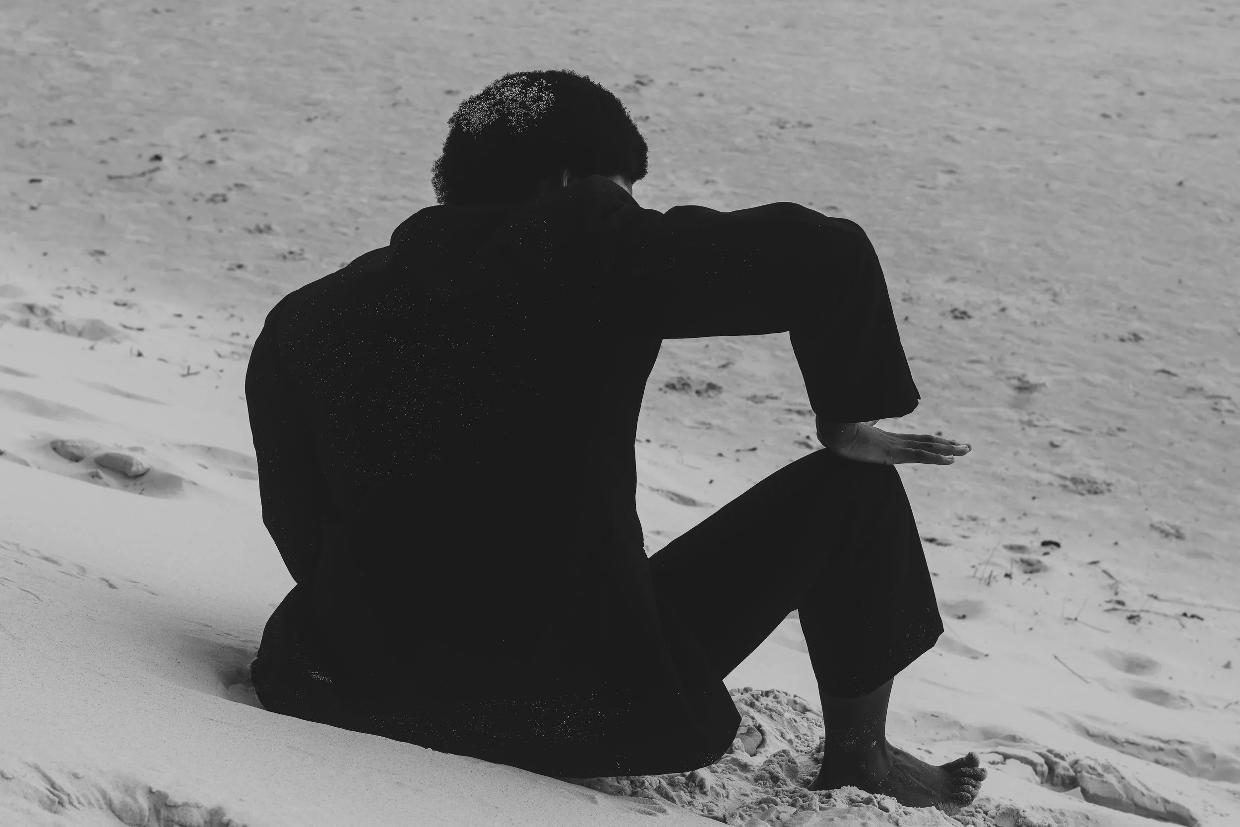 A person sitting on the sand at the beach, facing away, with one arm resting on their knee.