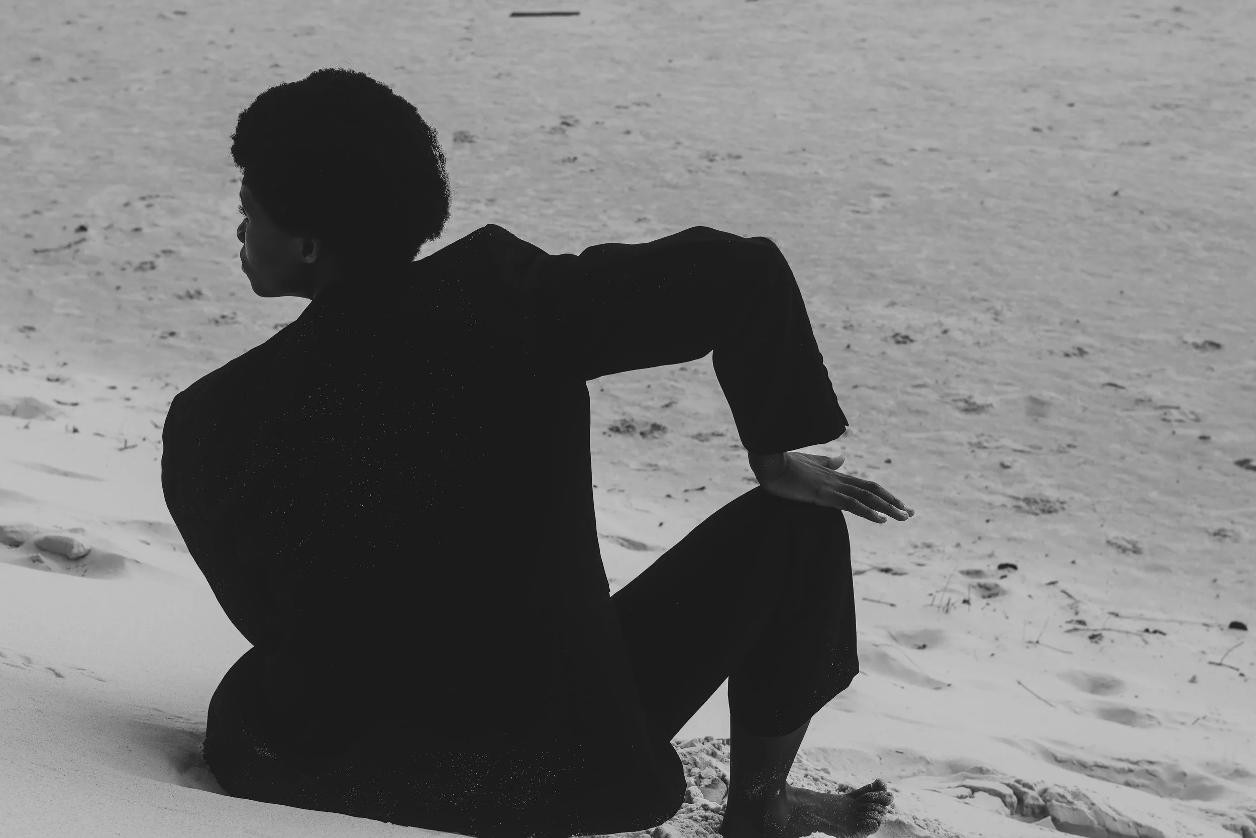 A person sitting on the sand at the beach, gazing into the distance, with their body slightly turned, wearing dark clothing.