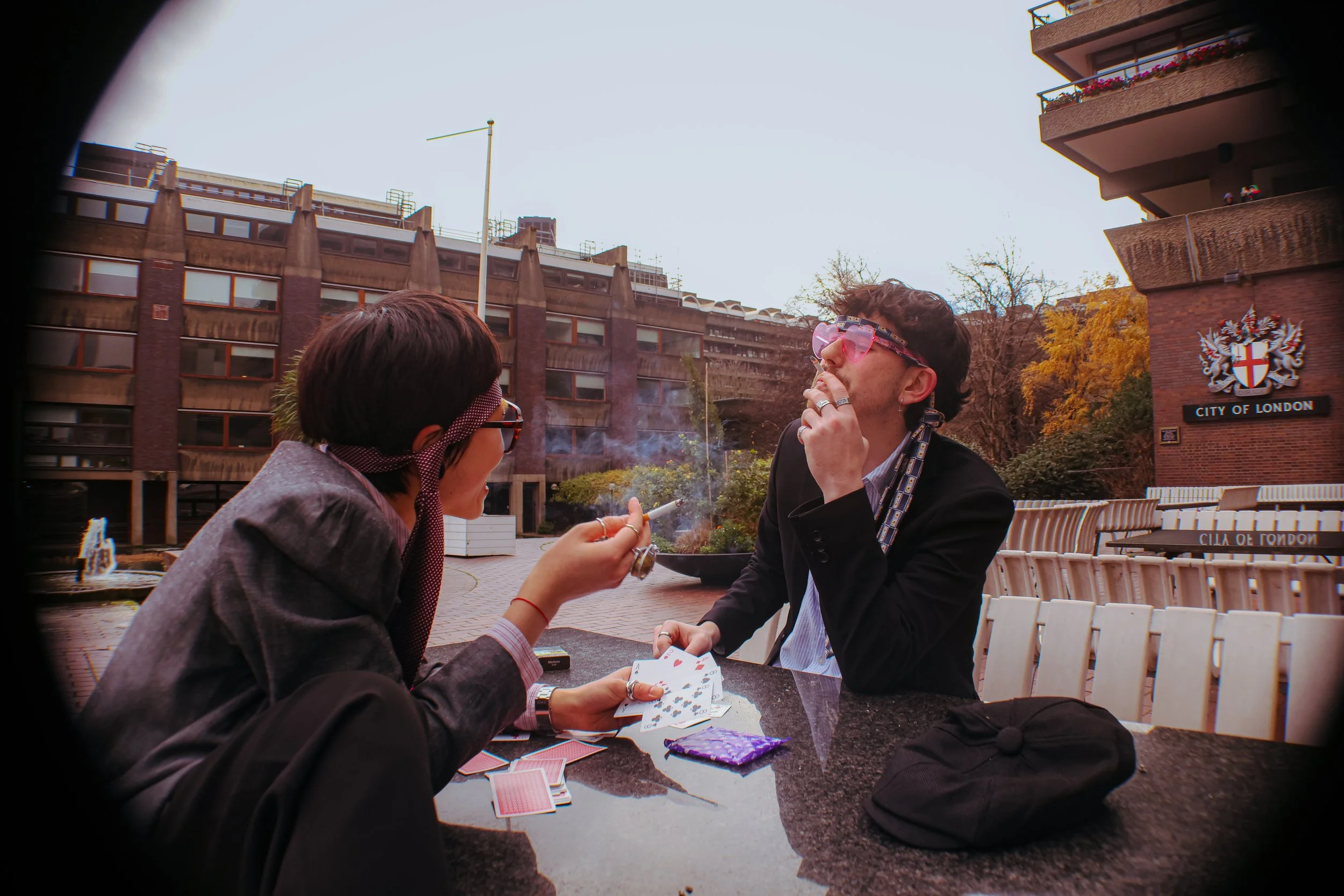 Two people playing cards outdoors at a table, with one smoking a cigarette and wearing heart-shaped sunglasses, in front of a city building with a "City of London" sign.