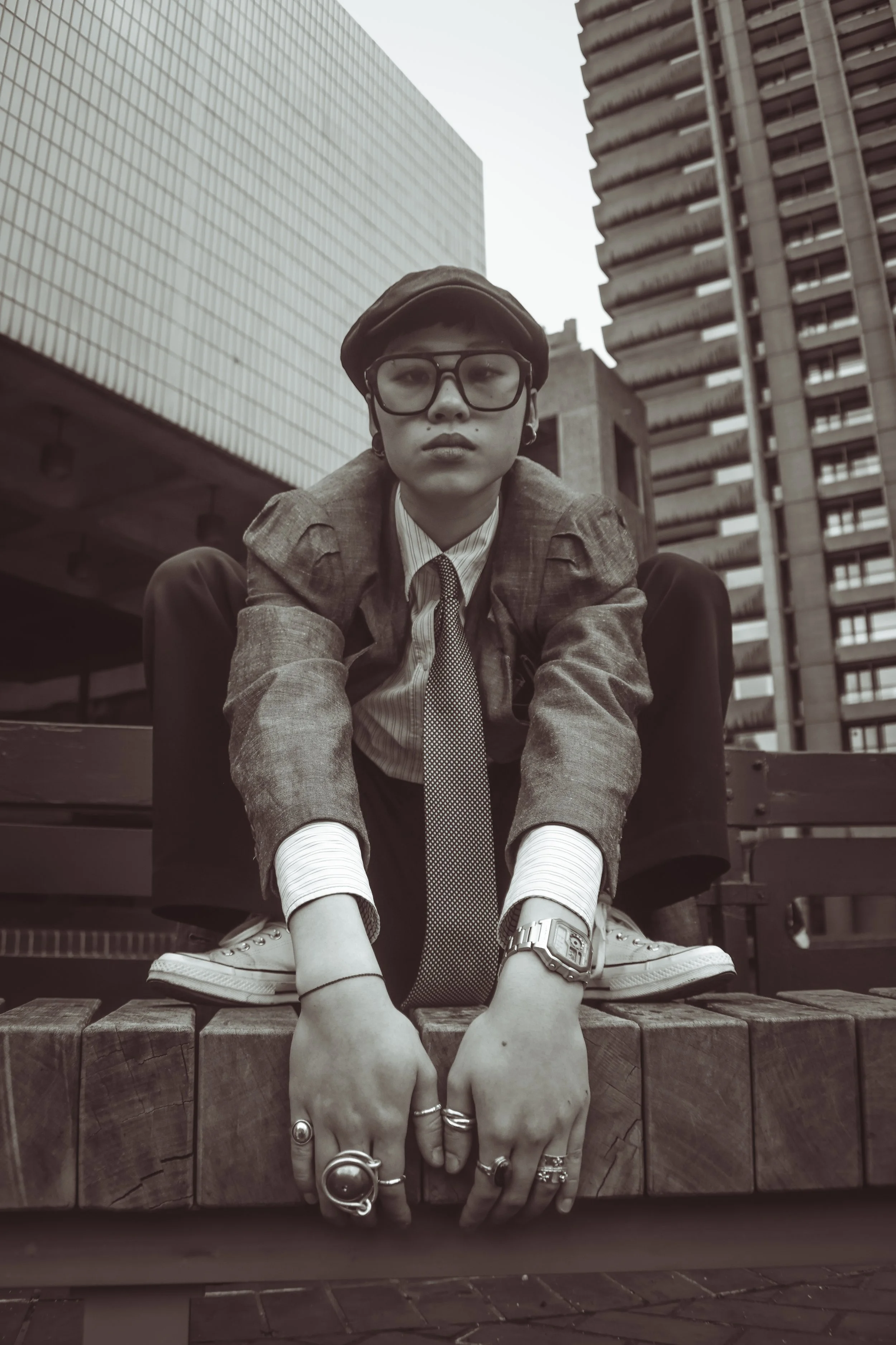 A young person with glasses, tattoos, and rings, dressed in a blazer, shirt, and tie, crouches on a wooden bench on a city street, with tall buildings in the background.