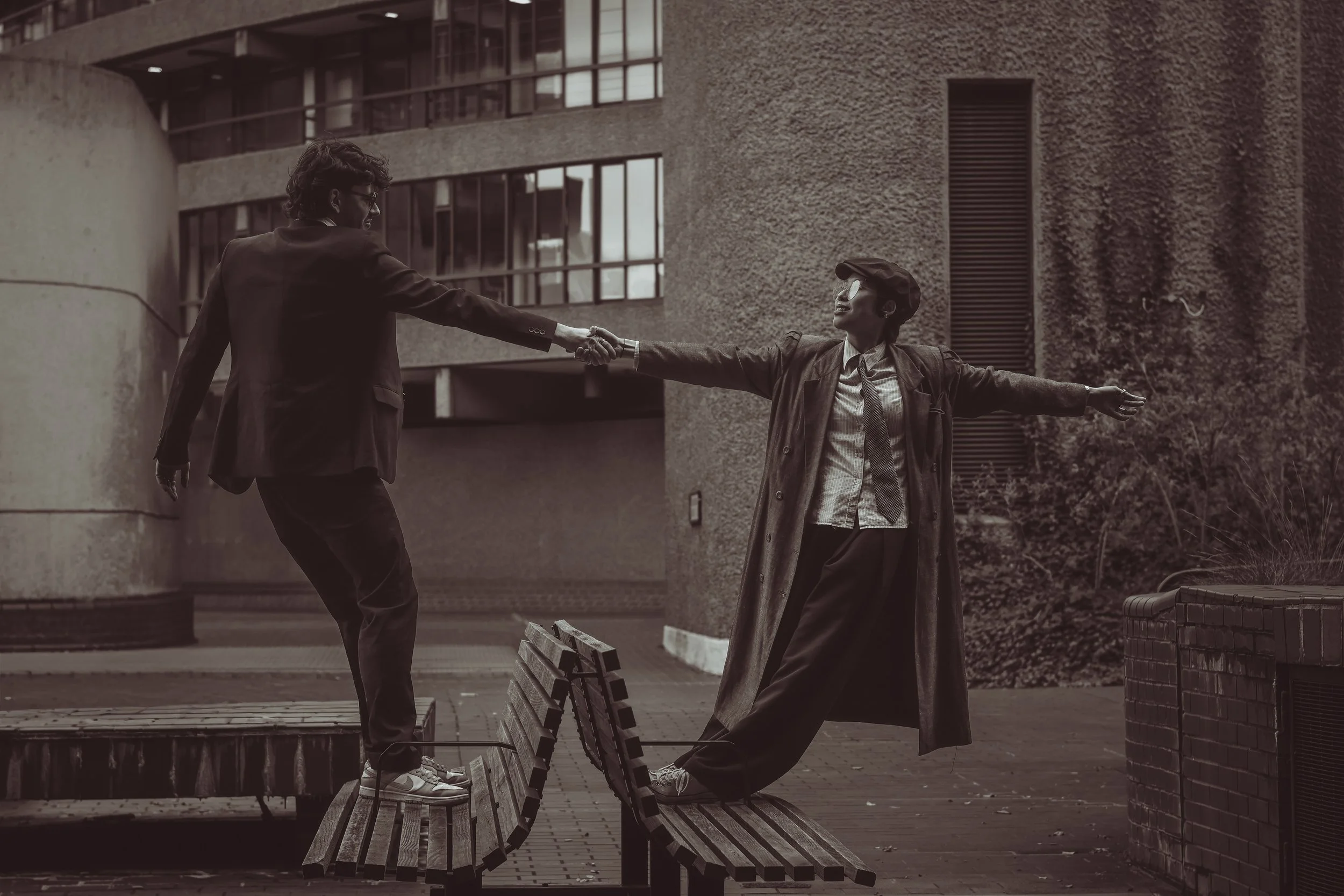 A black and white photo of a man and woman standing on a park bench. The man is standing on the seat of the bench, extending his hand to the woman. The woman is standing on the ground, reaching out to shake hands with the man, with her arms outstretched. Both are wearing coats. The background shows a large building with windows and some trees.