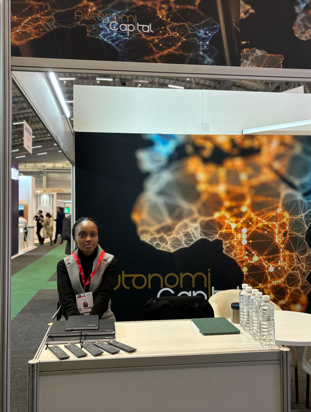A woman sitting behind a table in conference booth with a large black backdrop featuring a digital network and the words 'Autonomi Capital' at a trade show. Cape Town, South Africa.