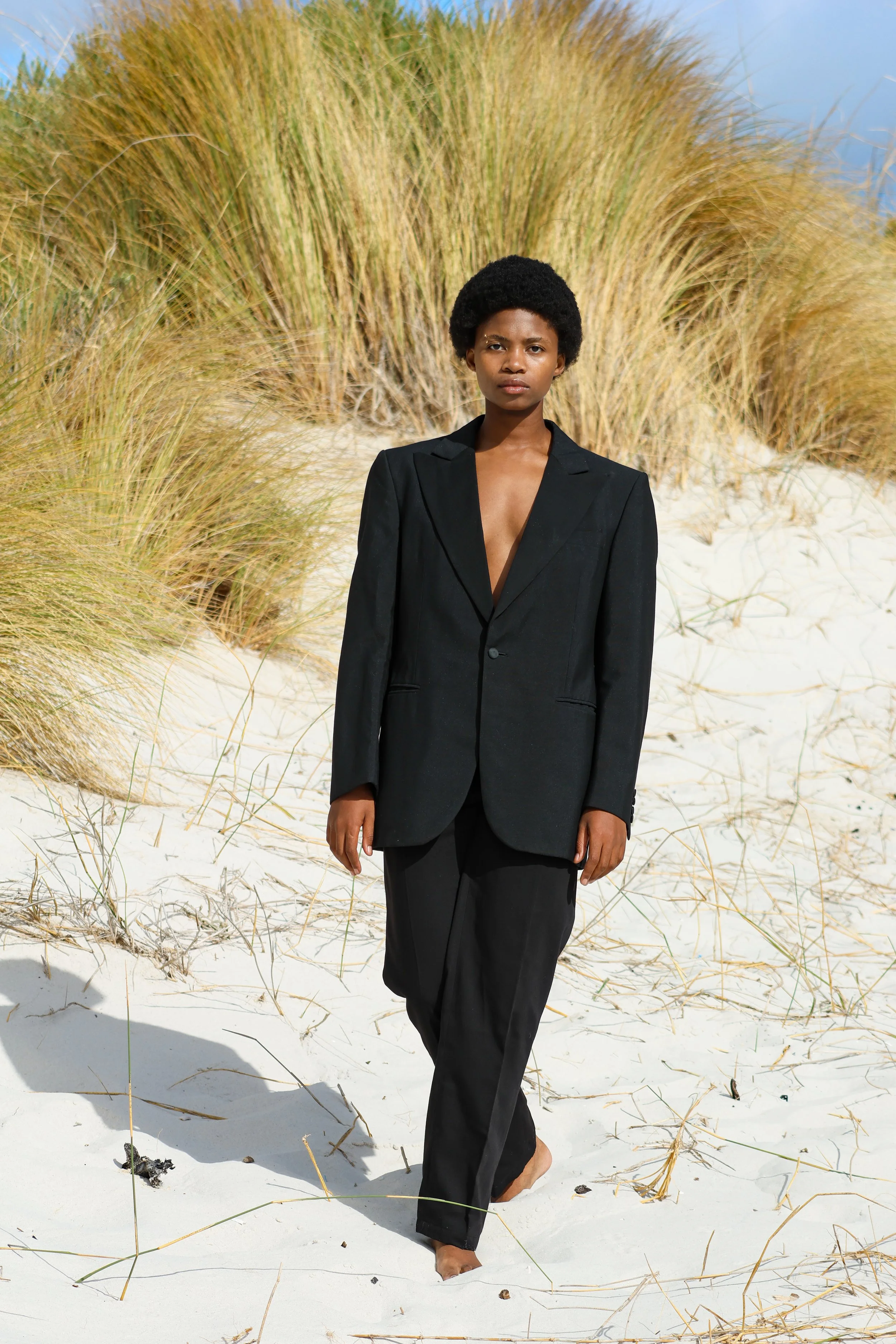A woman with a short afro hairstyle and dark skin wearing a black suit without a shirt underneath, walking barefoot on white sand near tall grasses. Noordhoek beach, Cape Town.