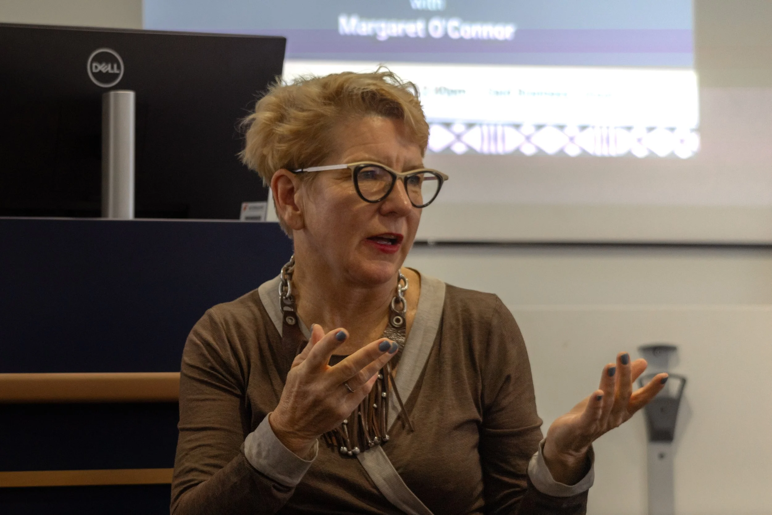 An older woman with short blonde hair and glasses, wearing a brown top and a chunky necklace, speaking and gesturing with her hands in front of a presentation screen and a Dell computer.