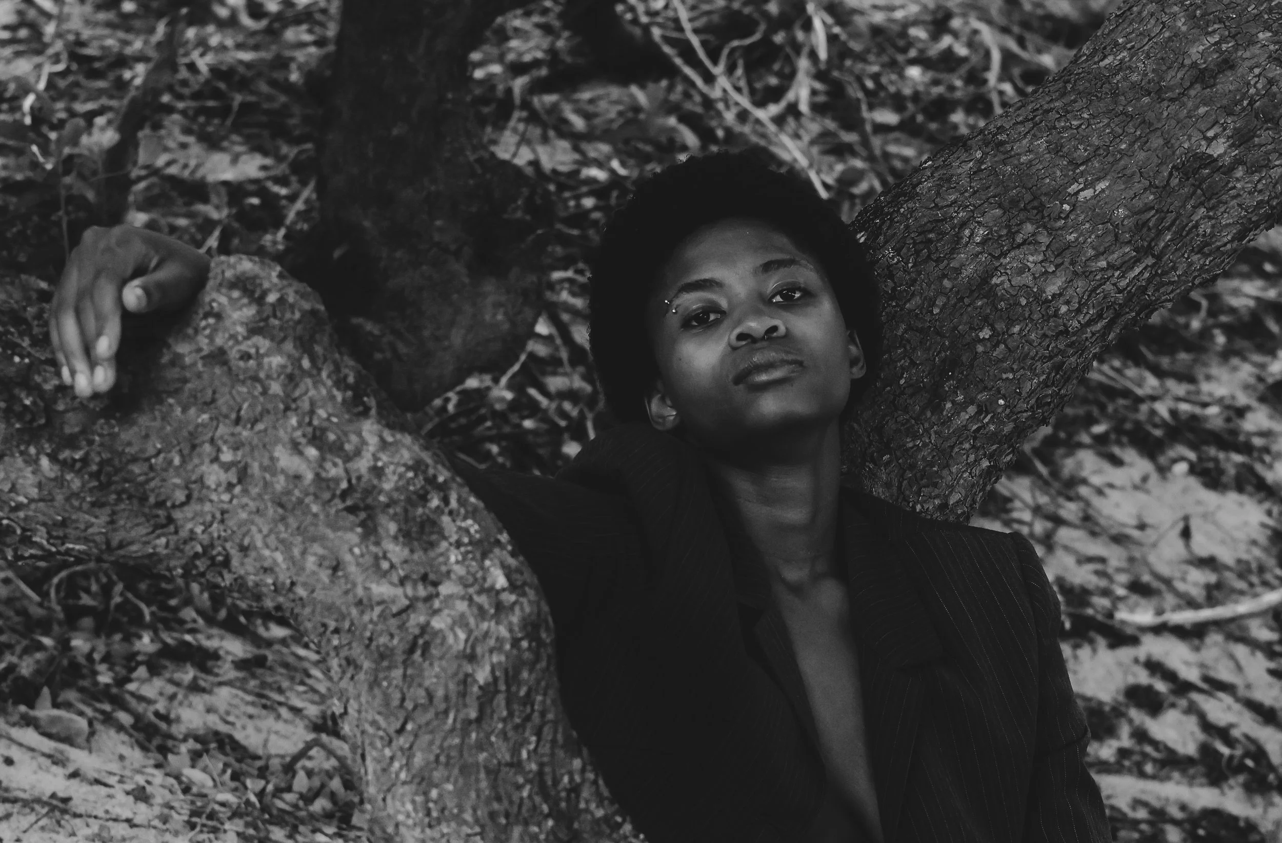 A portrait of a person with short curly hair and a confident expression leaning against a tree in a wooded outdoor setting, dressed in a dark blazer. Noordhoek beach, Cape Town.
