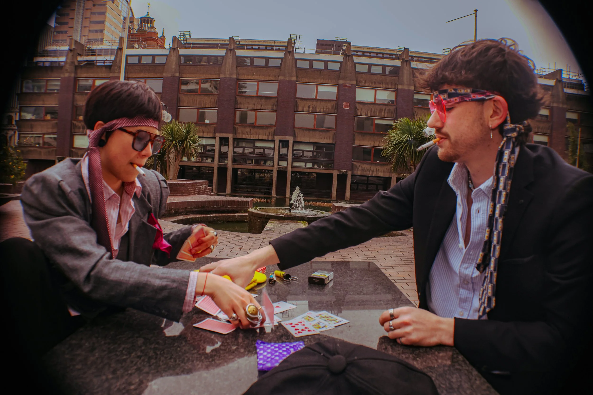 Two people with 80s style clothing and accessories playing cards outside on a table, with a modern building and fountain in the background.