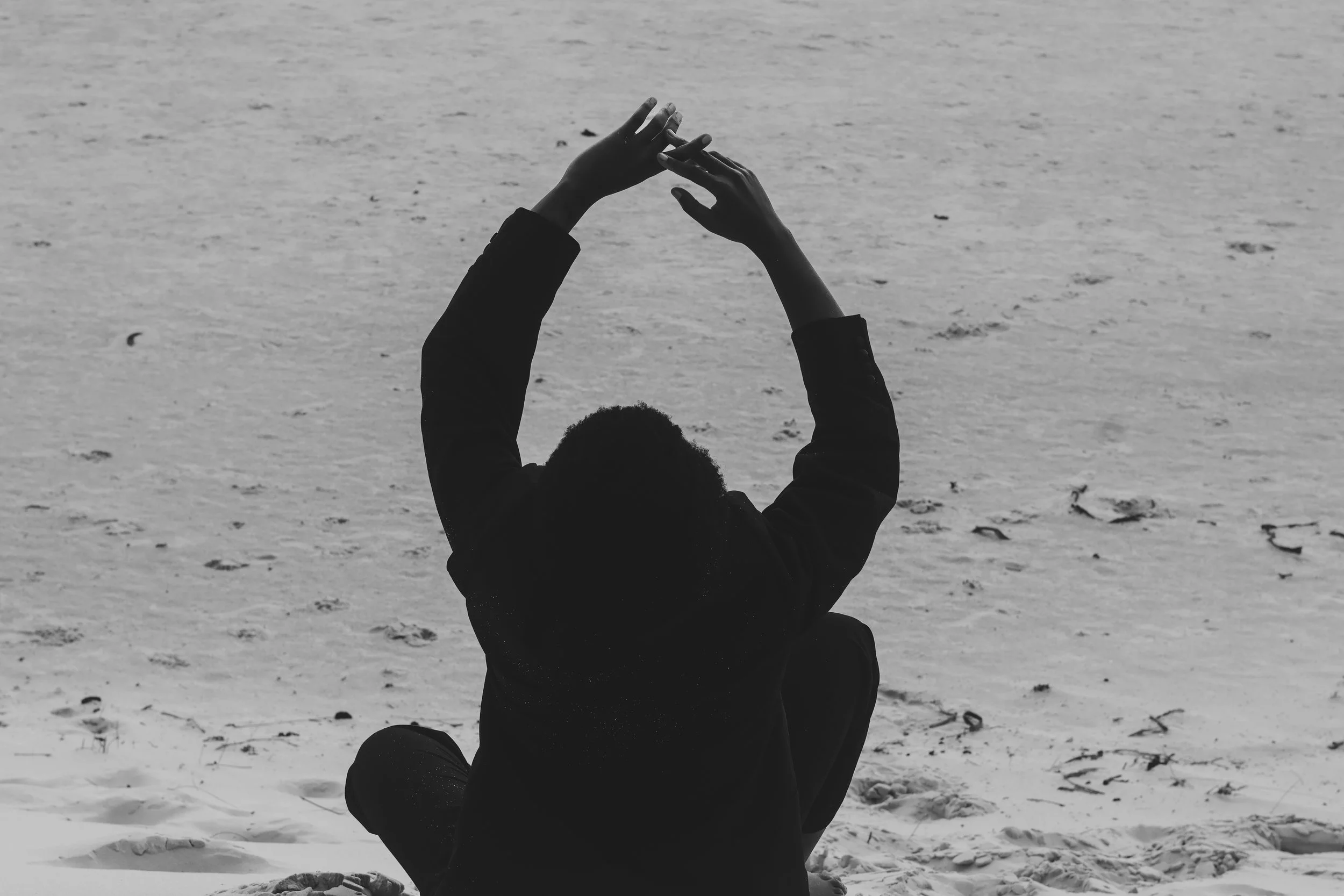 A person sitting cross-legged on the sand at the beach, facing away, with hands raised above their head in a yoga pose.