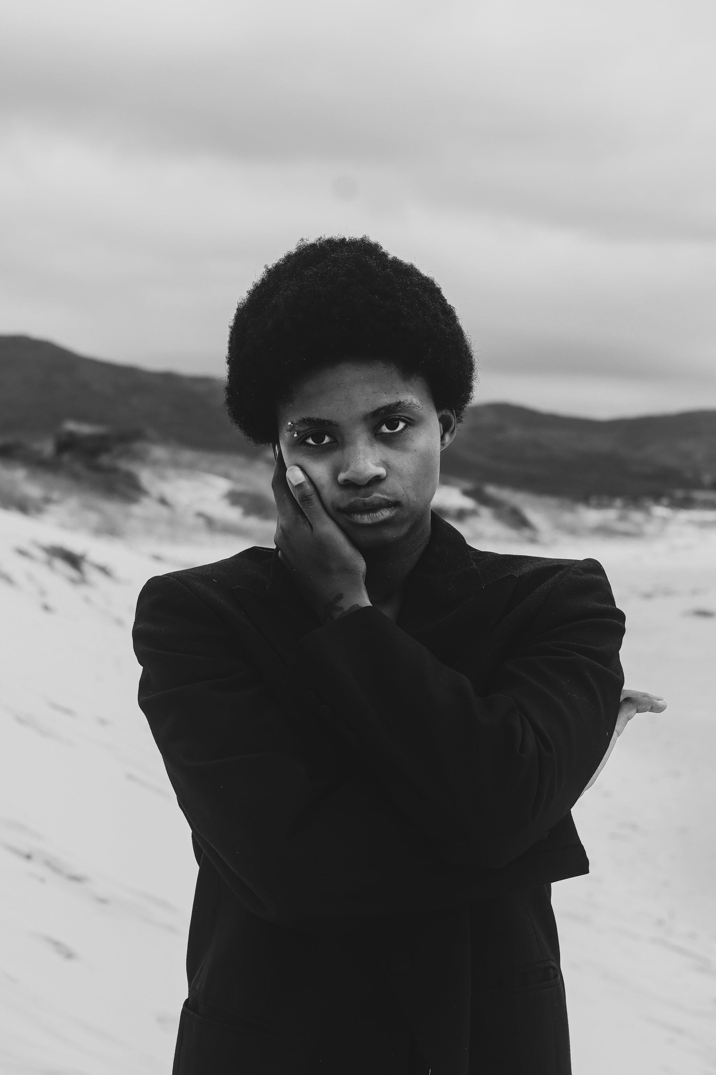 A black and white photo of a young woman with short curly hair, standing on a beach with sand dunes and hills in the background. She is wearing a dark coat, looking directly at the camera with her hand resting on her cheek. Noordhoek beach, Cape Town