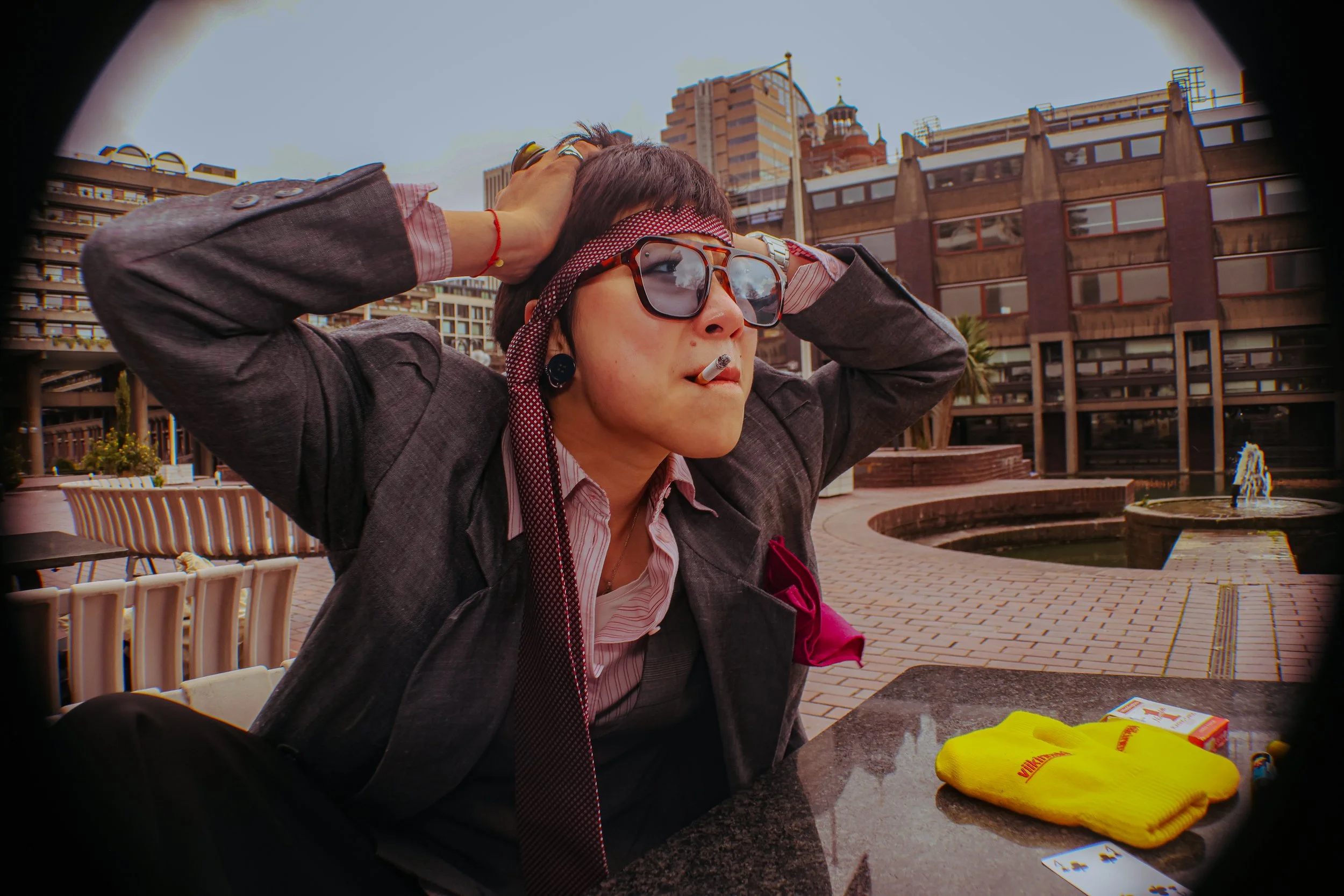 A woman wearing sunglasses, a headscarf, and a gray blazer sits at an outdoor table with her hands on her head, smoking a cigarette, in an urban setting with modern buildings and a fountain in the background.