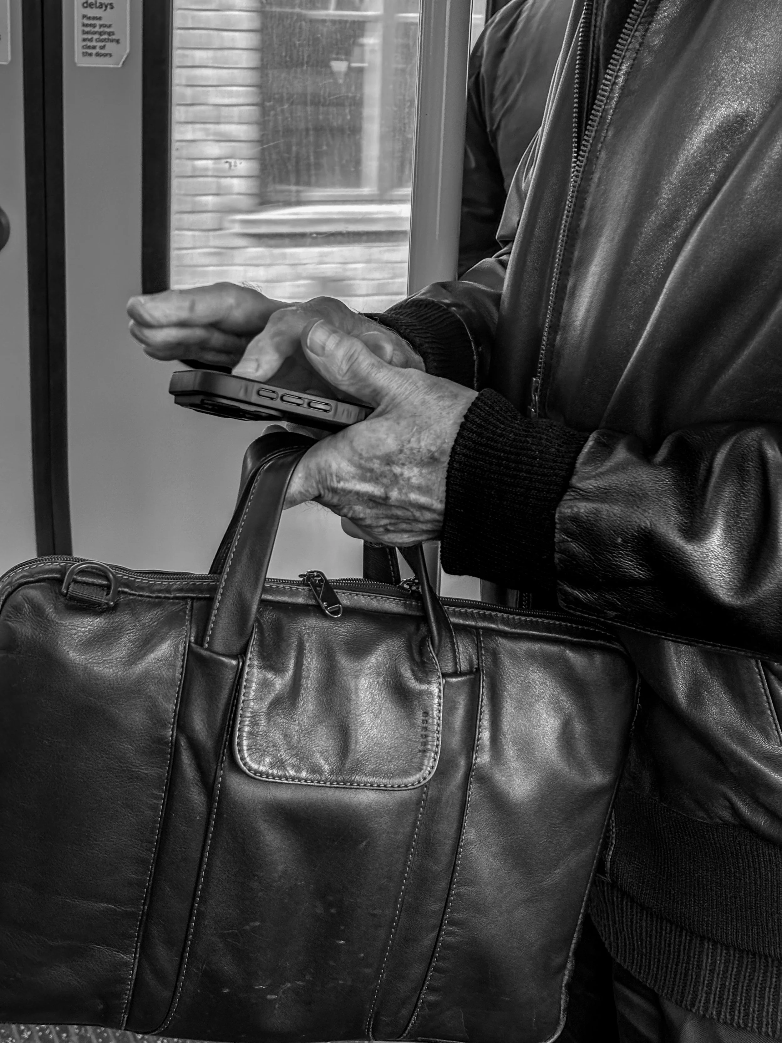 Close-up of a person wearing a leather jacket, holding a smartphone in their left hand and a large leather handbag in their right hand, inside a transit vehicle.
