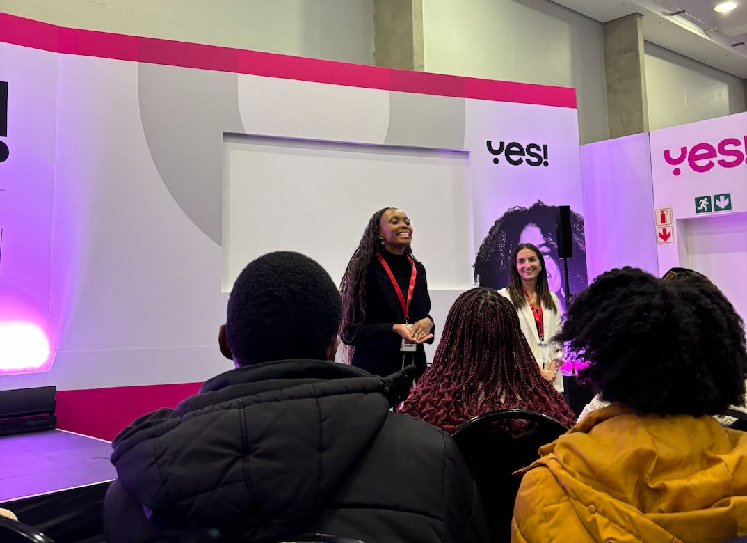 A woman speaking  on stage at YES! conference. Audience members are seated in the foreground, listening. The room has purple and pink lighting. Cape Town, South Africa.