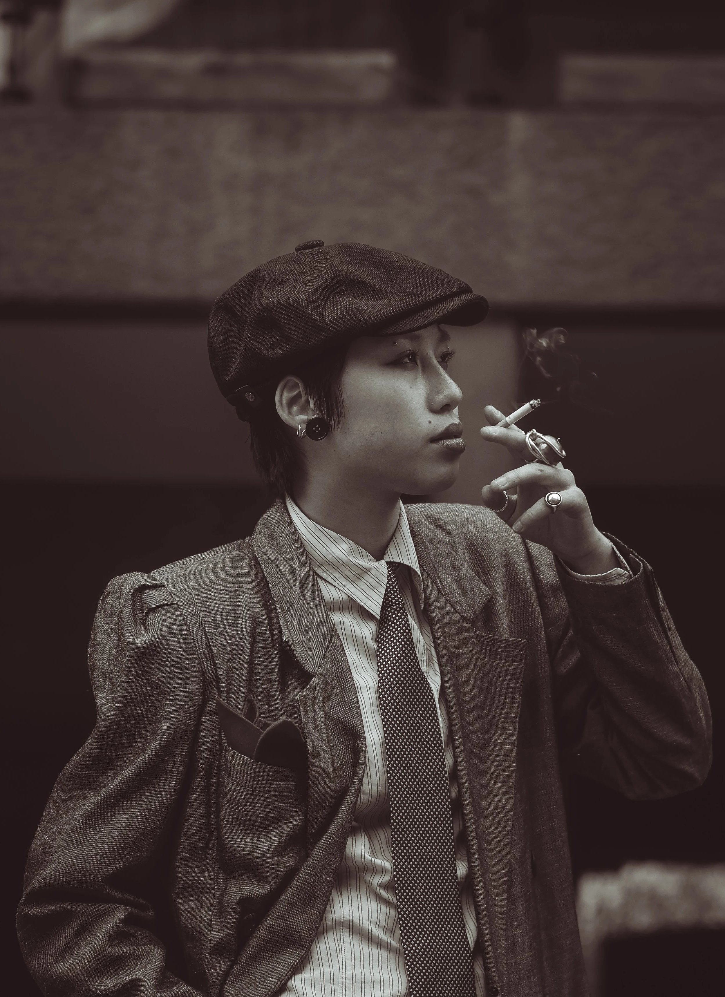 A woman with short hair and multiple earrings, wearing a blazer, a striped shirt, and a tie, is smoking a cigarette. She is also wearing a cap and has rings on her fingers.