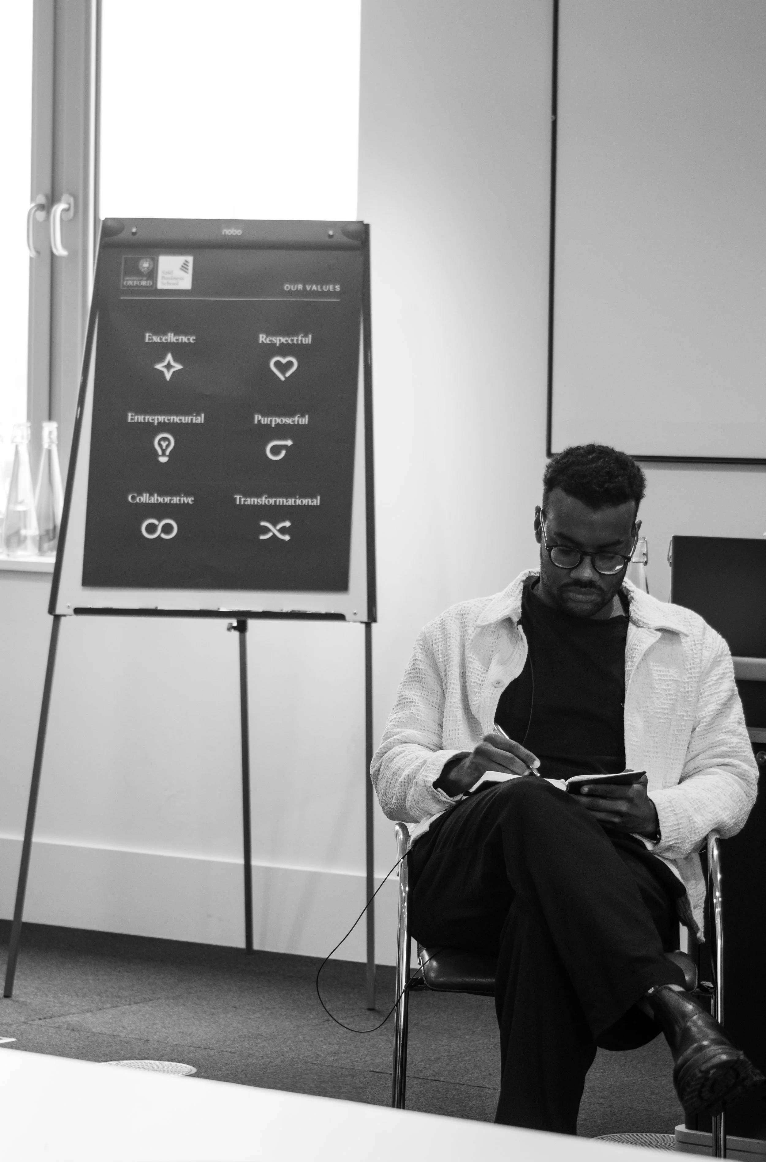 A man with glasses sits in a chair, writing in a notebook, with a presentation board in the background displaying values such as excellence, respectful, entrepreneurial, purposeful, collaborative, and transformational.