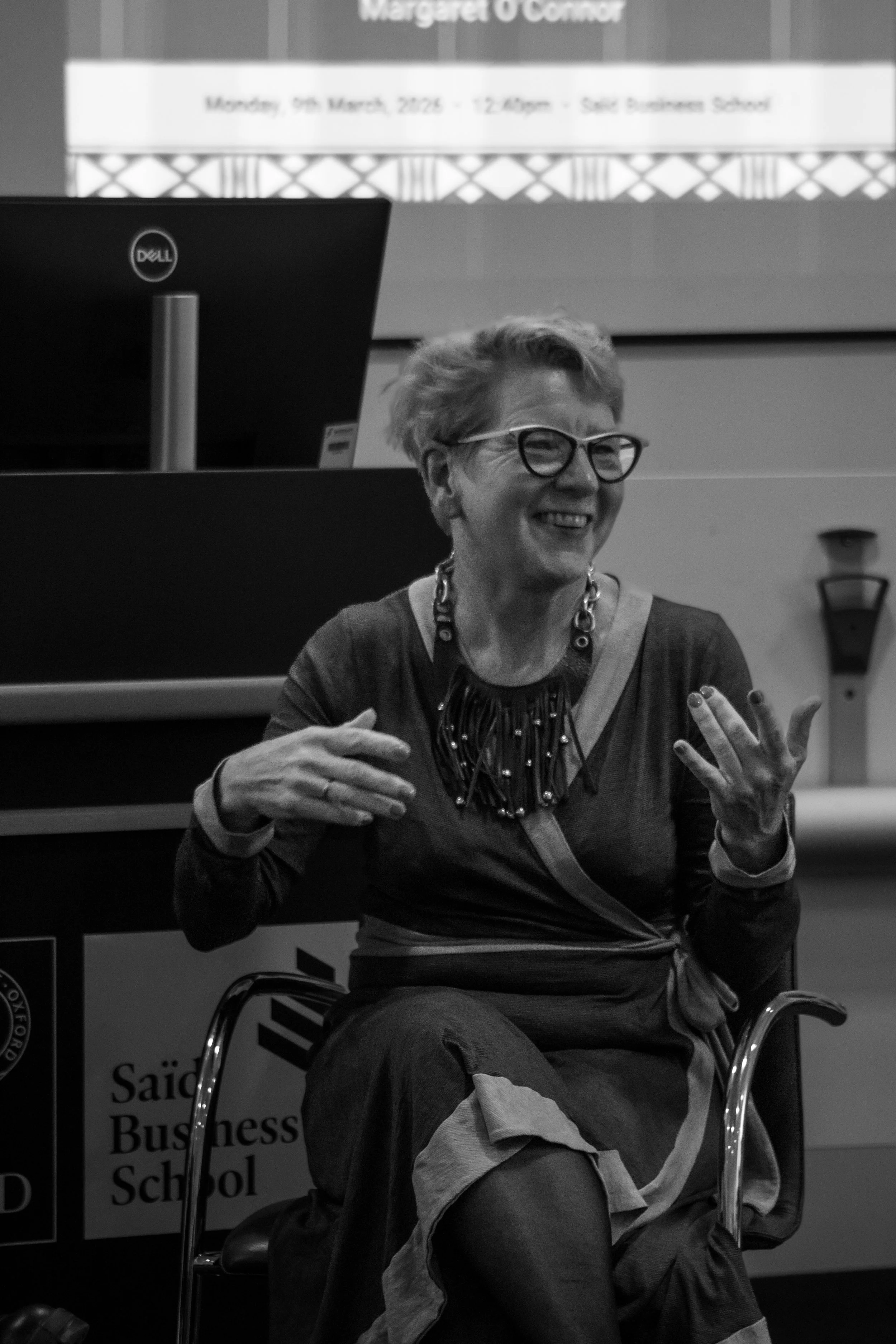A balck and white portrait of a woman with short, wavy hair and glasses, smiling and gesturing with her hands, sitting in a chair, giving a talk at the Oxford Edward Saïd Business School.