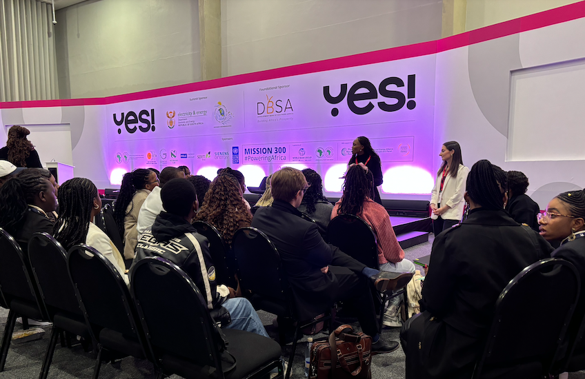 Two women speaking a conference with a stage and backdrop displaying 'YES!' and various logos, including 'DUBSA' and messages like 'Mission 300 #poweringAfrica'. Cape Town, South Africa.