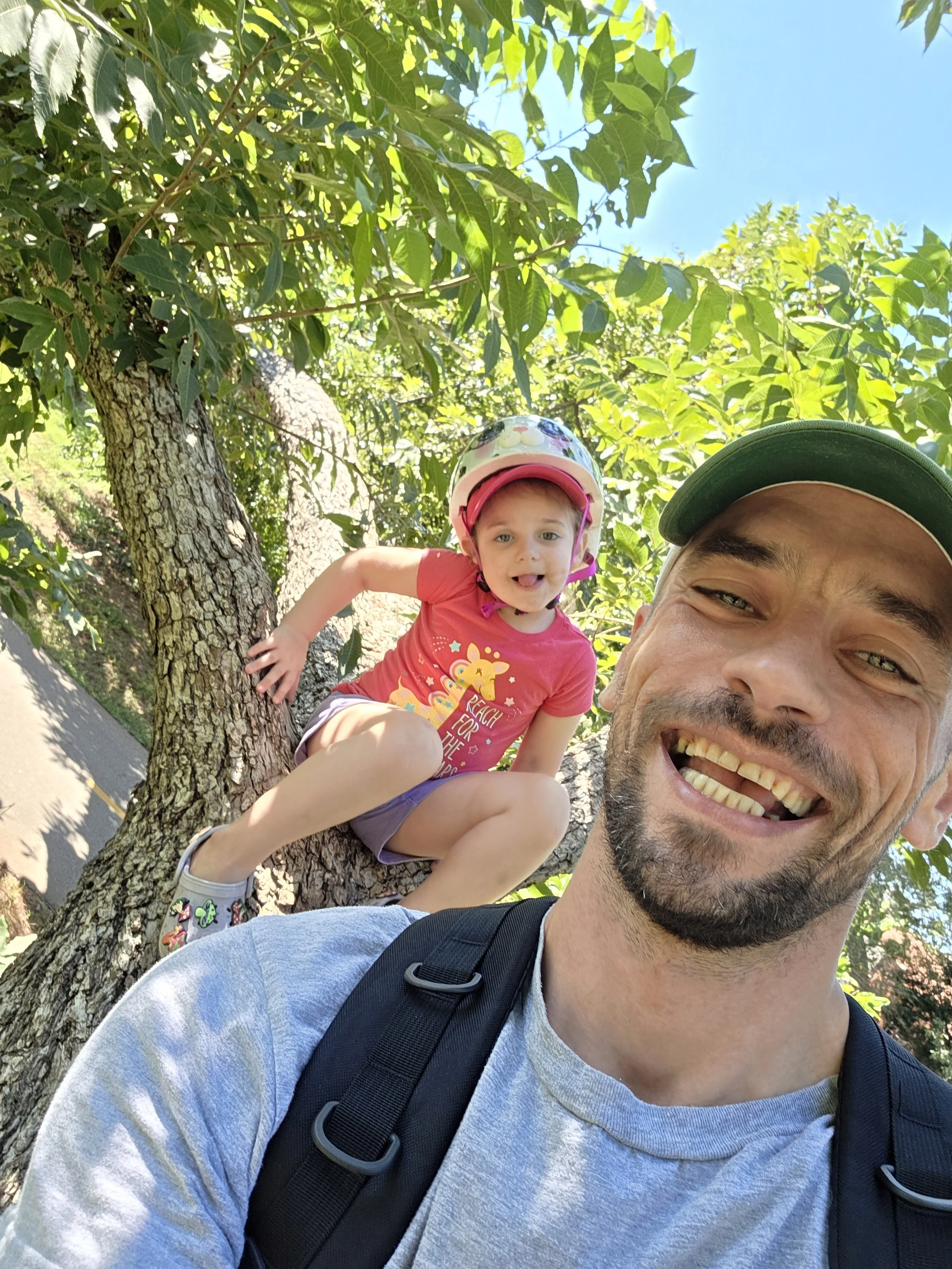 A smiling man and a young girl wearing a helmet are outdoors near a tree, with a clear blue sky in the background.