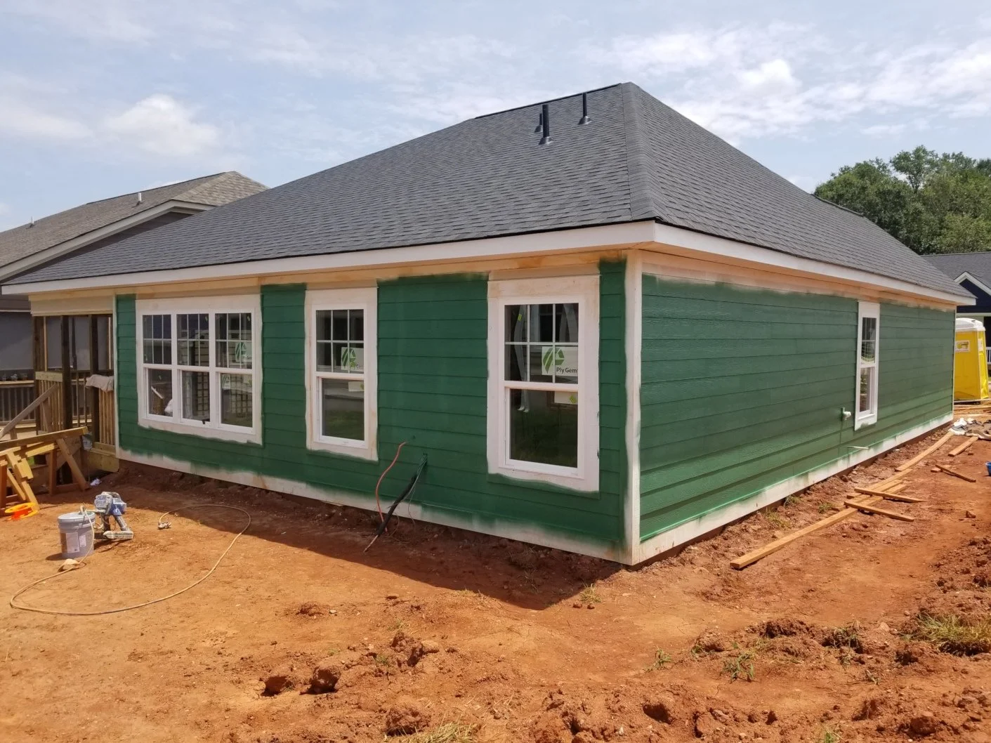 Newly constructed house with green exterior siding, white window frames, and a gray shingle roof on a dirt lot with construction tools and materials around.