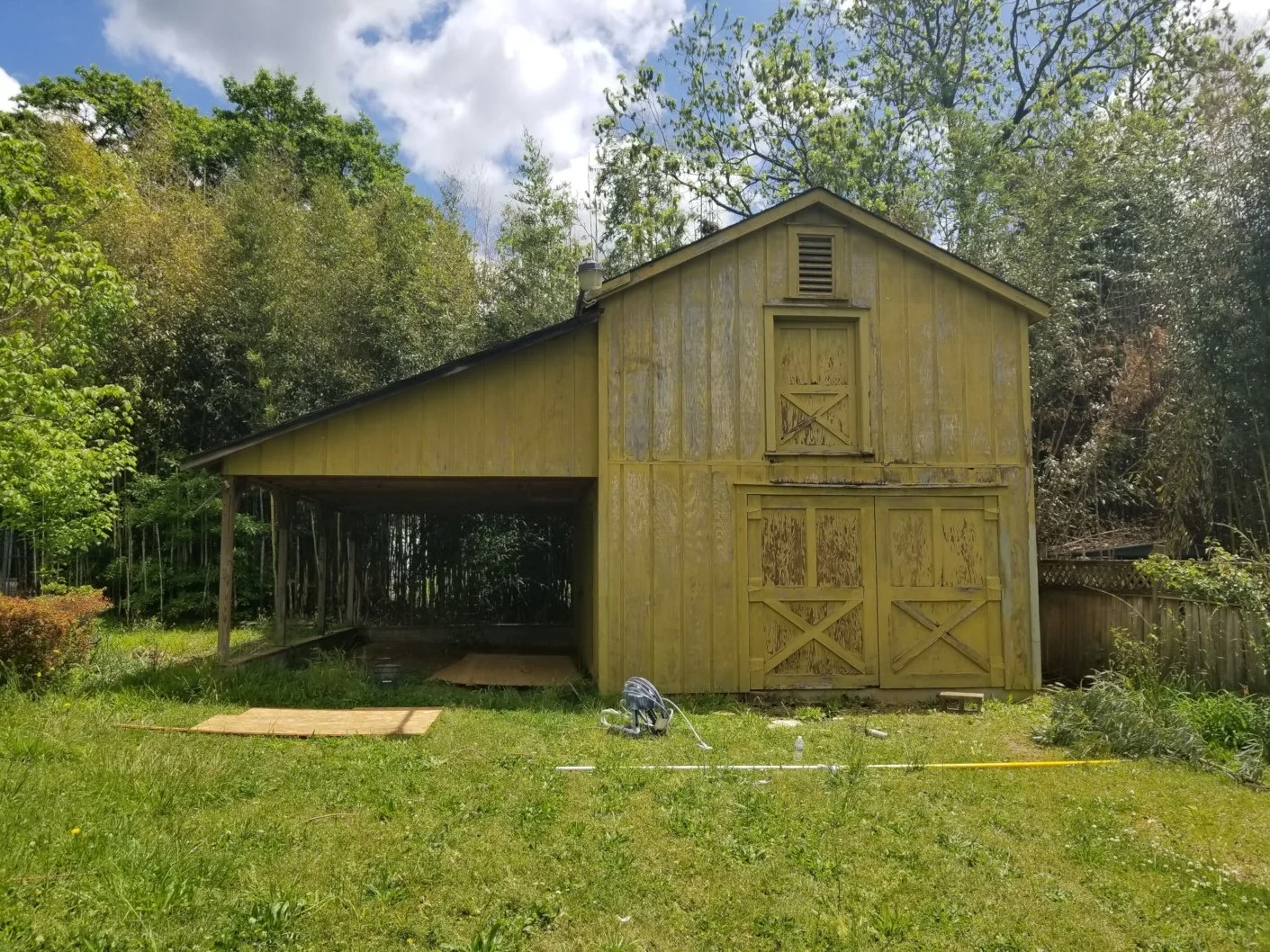 A partially renovated wooden yellow barn in a grassy backyard with trees in the background and partly cloudy sky.