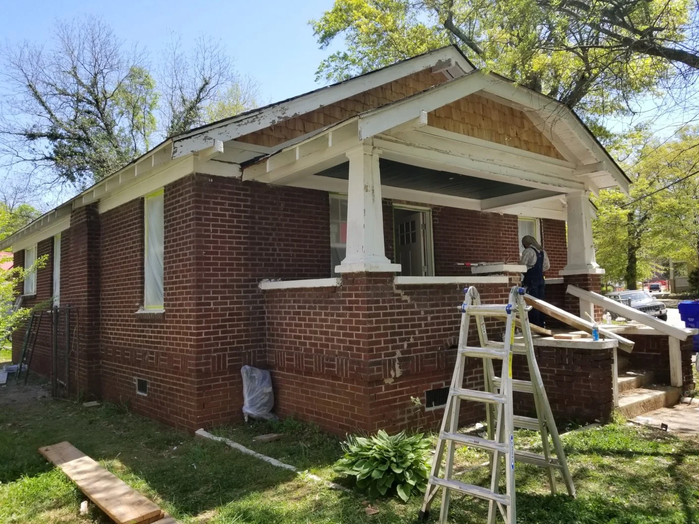 Two workers are renovating the porch of a brick house, with a ladder and construction materials around, and trees in the background.
