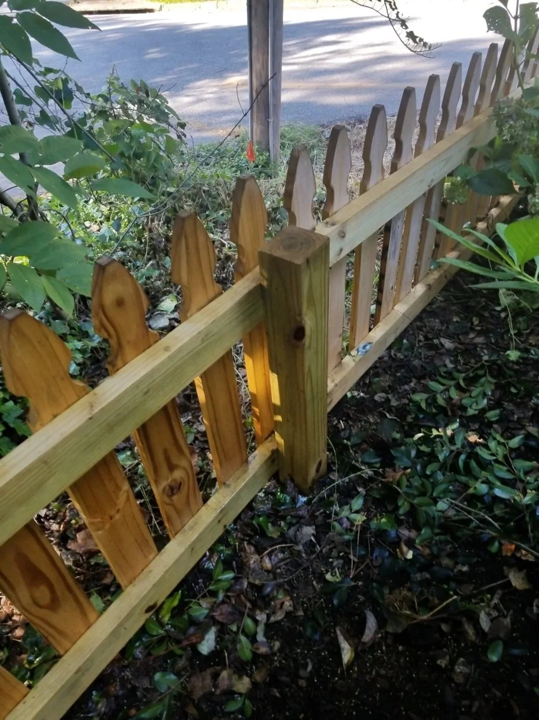 New wooden picket fence installed along the ground, with some greenery and bushes nearby, and a street visible in the background.